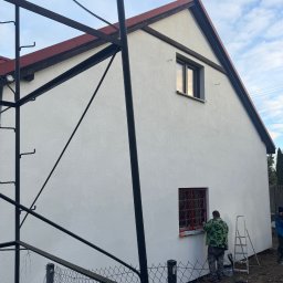 USŁUGI REMONTOWO BUDOWLANE - Pracownik w Czersku maluje czerwoną farbą ramę okienną na tle świeżo otynkowanej, białej elewacji domu, widoczne rusztowanie i drabina.