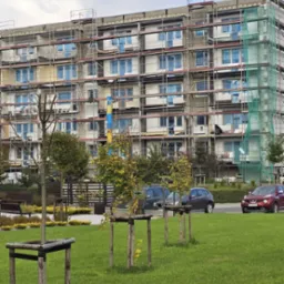 Wielopiętrowy budynek w trakcie ocieplania, osłonięty rusztowaniami i siatką ochronną. Okna zabezpieczone folią. Na pierwszym planie trawnik z młodymi drzewkami.