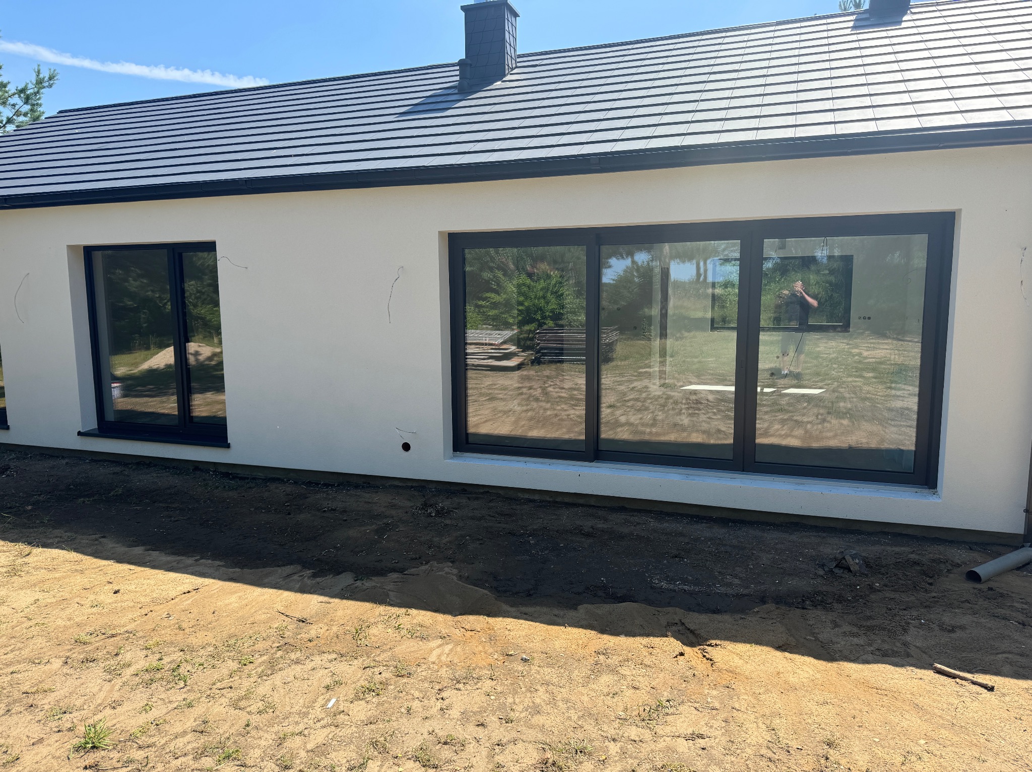 Nowoczesny dom jednorodzinny z jasną elewacją i dużymi oknami z czarnymi ramami, widoczny fragment dachu z ciemnej dachówki oraz teren wokół budynku w trakcie prac wykończeniowych.