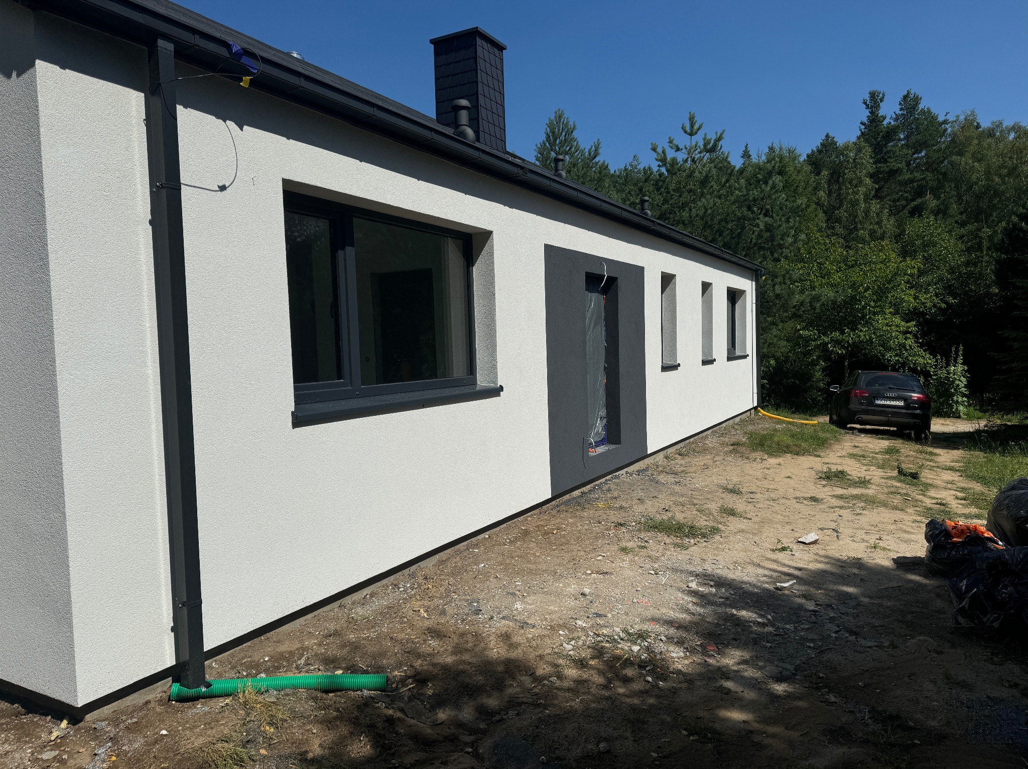 Nowoczesny dom jednorodzinny z białą elewacją i antracytowymi oknami, wykończony z dbałością o detale, widok z boku na tle zieleni, okno częściowo zabezpieczone folią ochronną.