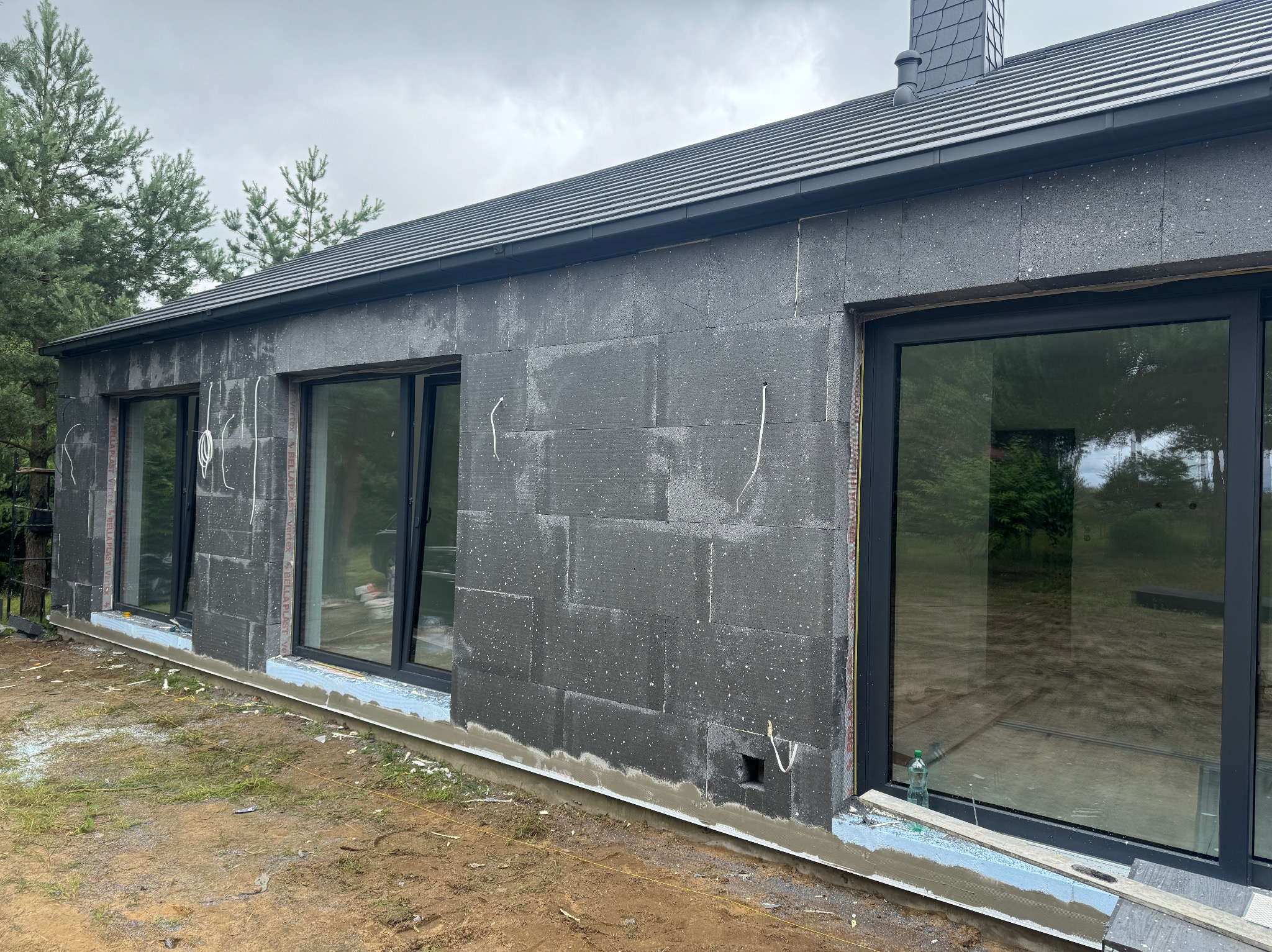 Dom w trakcie izolacji: ściany pokryte ciemnoszarymi płytami styropianowymi, widoczne okna z czarnymi ramami, fragment dachu z szarej dachówki, teren wokół domu częściowo zagospodarowany.