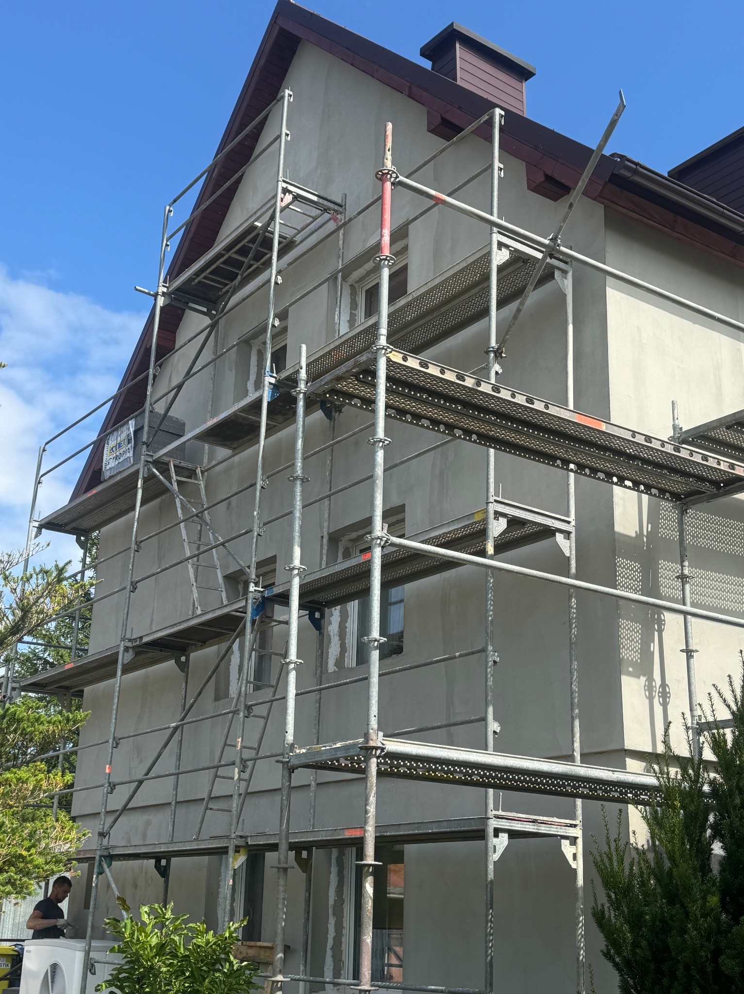 Dom jednorodzinny w trakcie nakładania szarego tynku, widoczne rusztowania oraz fragment dachu z brązową dachówką.