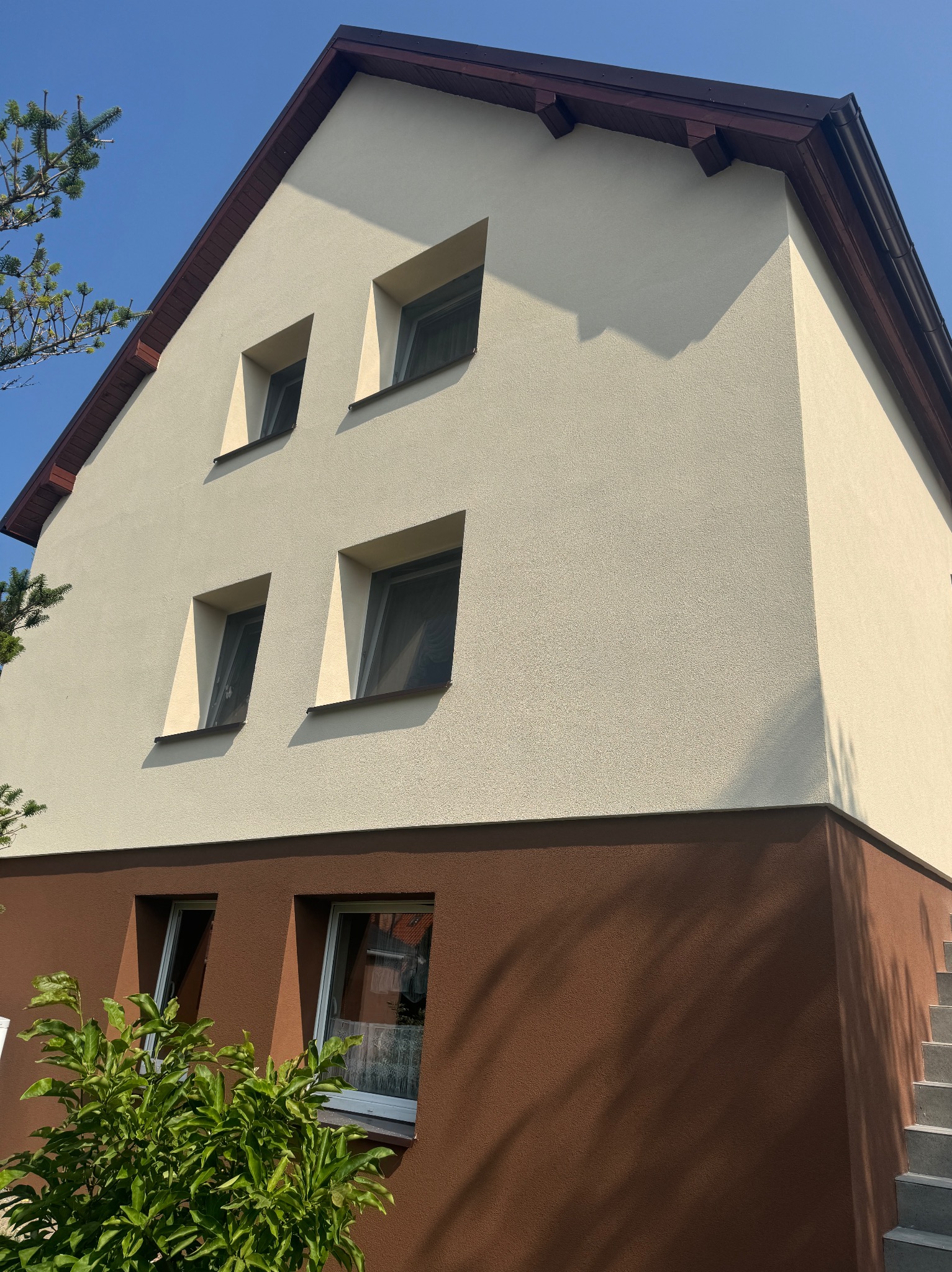 Dwukondygnacyjny dom z jasnobeżową elewacją górnej części i brązową dolnej, widoczne cztery okna na górze i dwa na dole, brązowe obramowanie dachu i okien, oraz fragment schodów i zieleni.