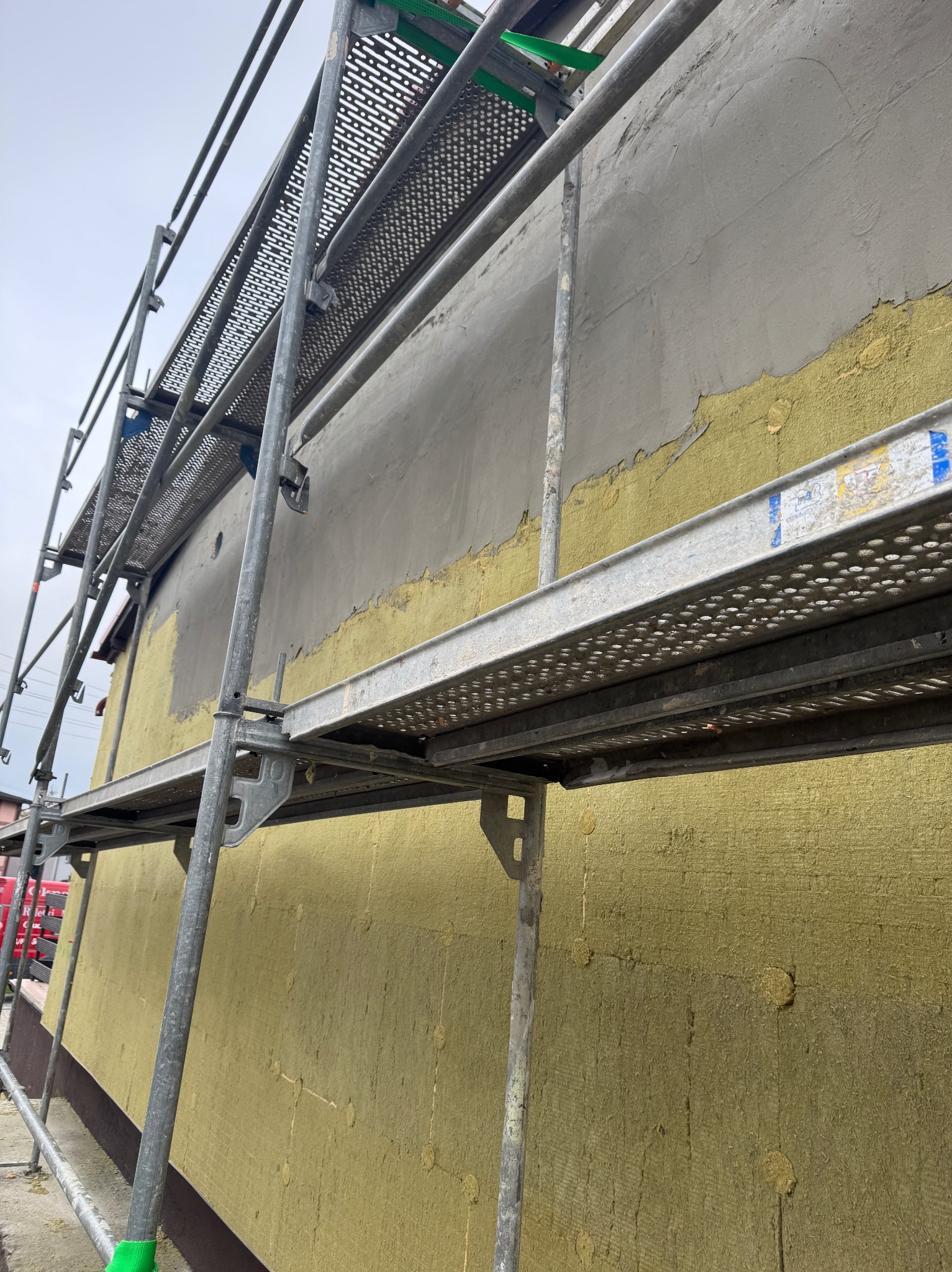 Elewacja budynku w trakcie prac: widoczna warstwa izolacyjna z wełny mineralnej, częściowo pokryta szarą warstwą tynku, rusztowanie z metalowymi pomostami.