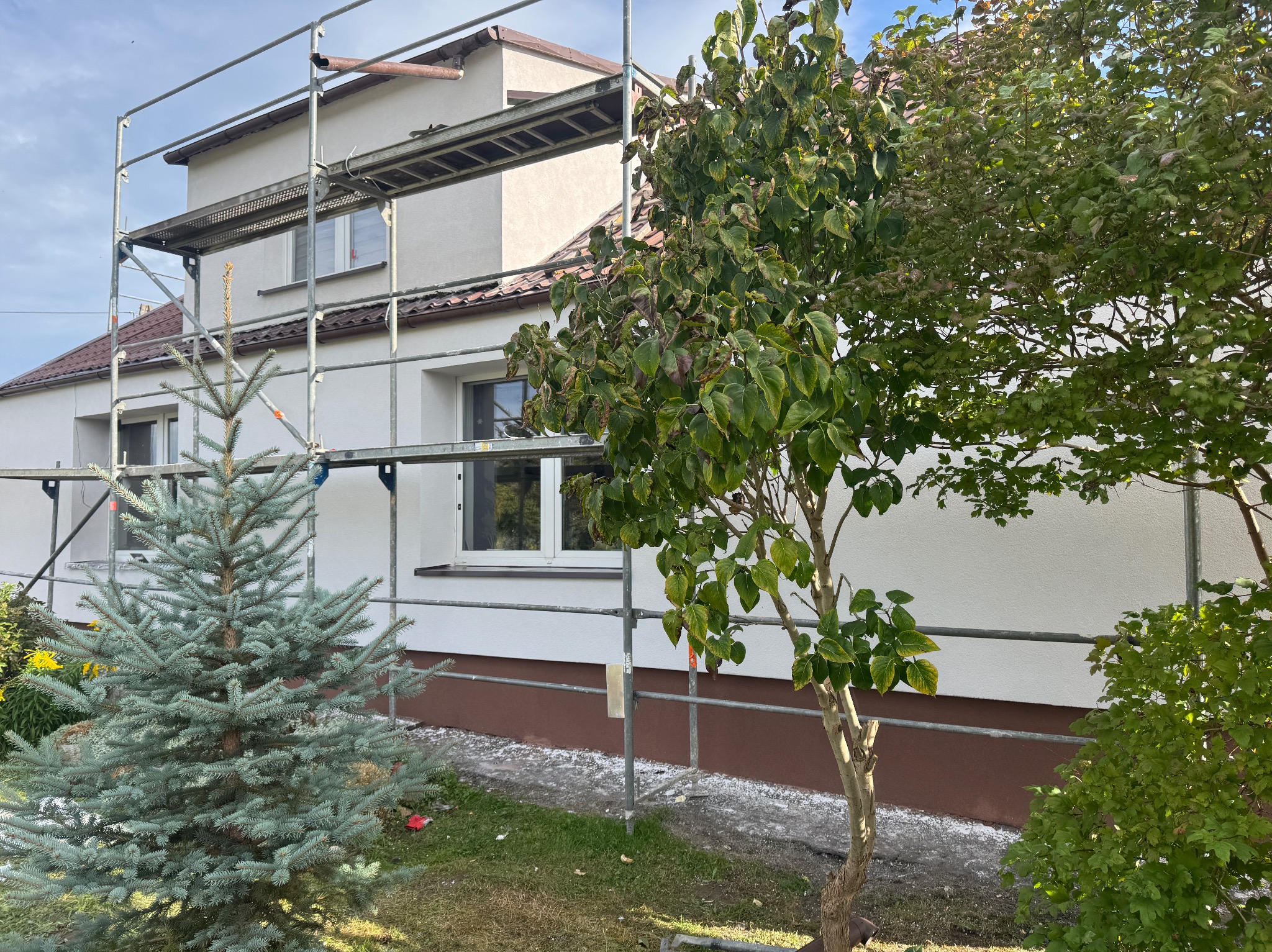 Dom jednorodzinny z białymi ścianami i brązowym cokołem w trakcie prac elewacyjnych, widoczne rusztowania i zieleń drzew