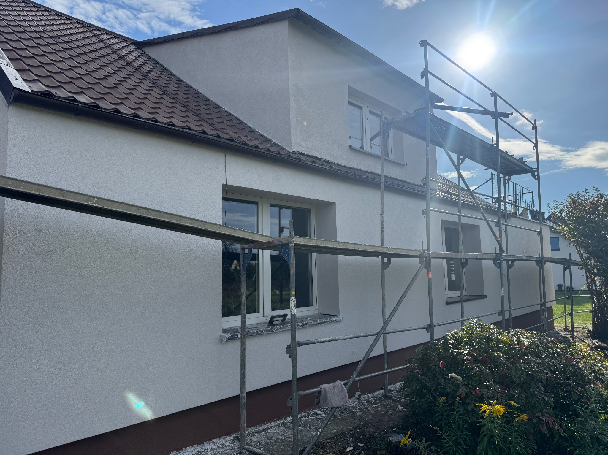 Dom jednorodzinny w trakcie prac elewacyjnych: świeżo otynkowana, jasnoszara ściana z widocznymi rusztowaniami, zabezpieczonymi oknami i fragmentem brązowego cokołu. Słoneczny dzień.