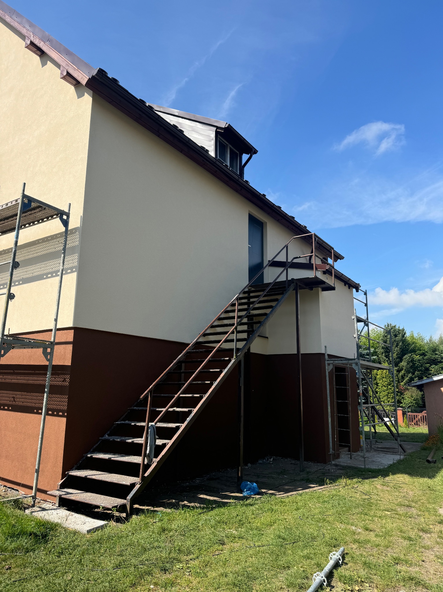 Dwukondygnacyjny budynek z beżowo-brązową elewacją, metalowe schody zewnętrzne prowadzące do drzwi na piętrze, rusztowania przy ścianach, błękitne niebo w tle.
