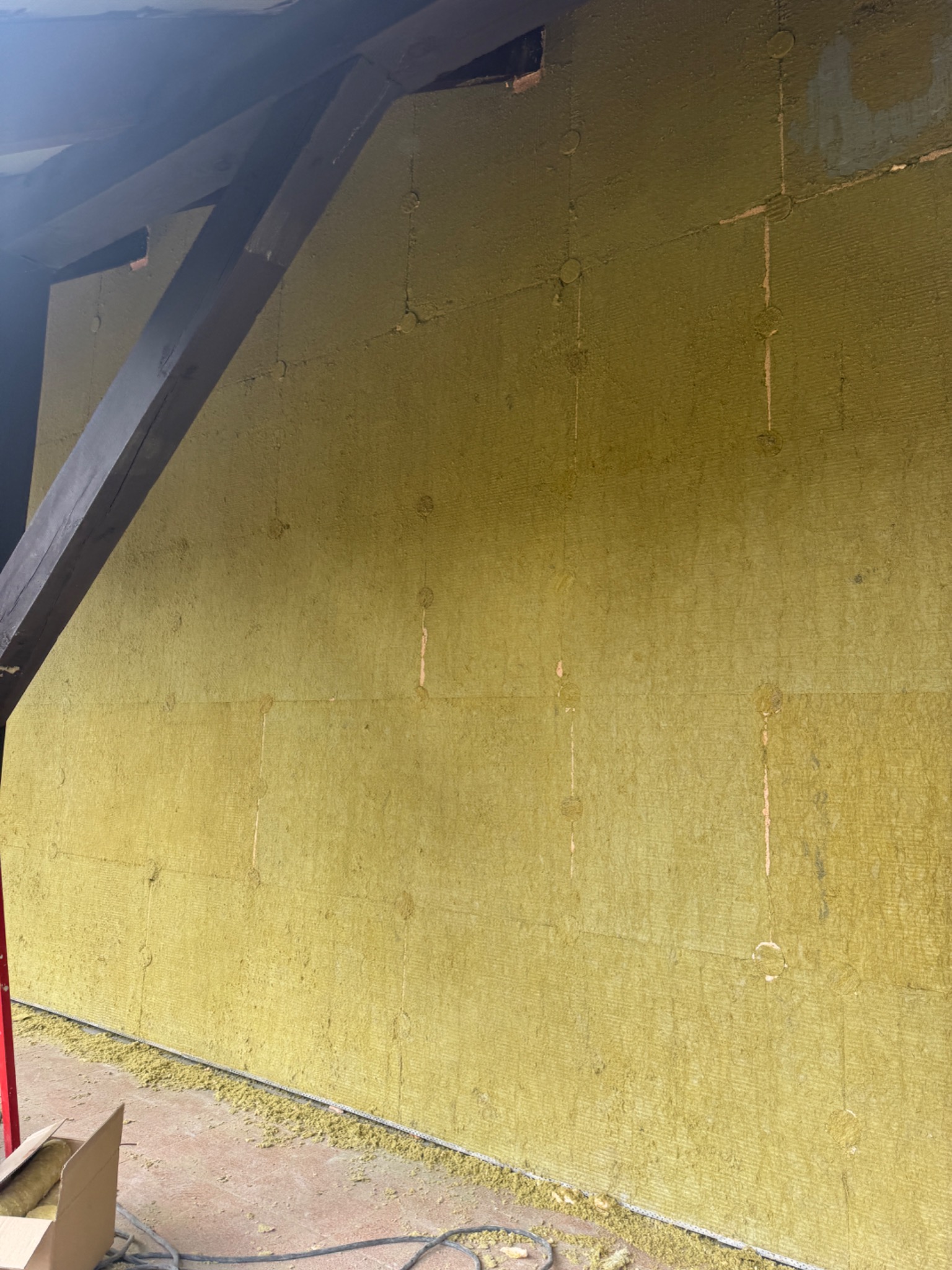 Ściana budynku w trakcie ocieplania wełną mineralną, widoczne kołki mocujące i fragment drewnianej konstrukcji dachu.