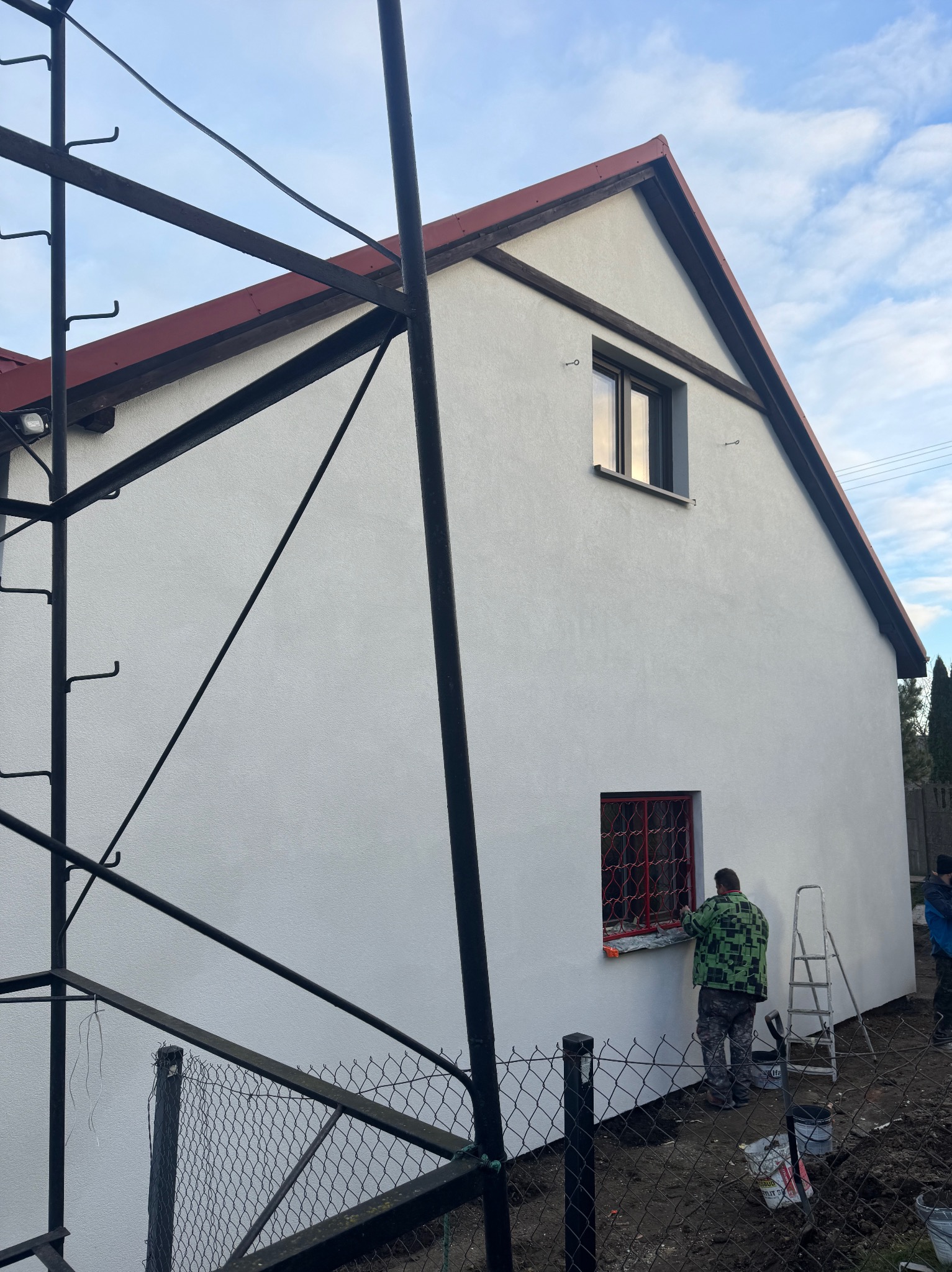 Pracownik w Czersku maluje czerwoną farbą ramę okienną na tle świeżo otynkowanej, białej elewacji domu, widoczne rusztowanie i drabina.
