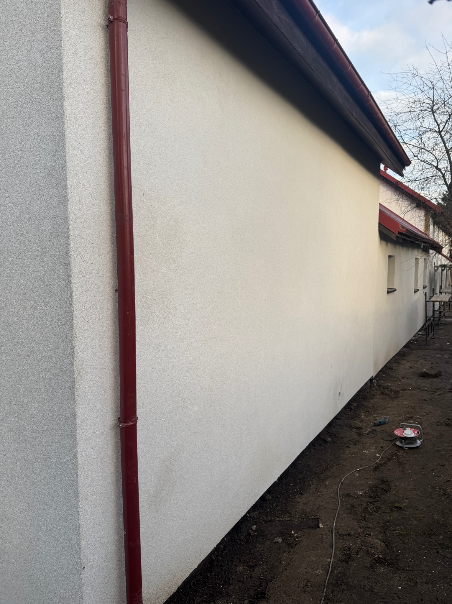 Świeżo otynkowana elewacja budynku z czerwoną rynną spustową i rozłożonym przedłużaczem na ziemi, widok z boku.