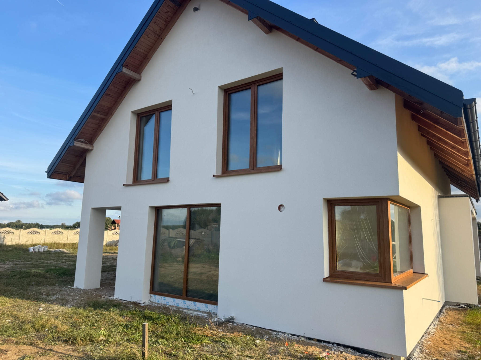 Biała elewacja domu jednorodzinnego z drewnianymi oknami i wykończeniem dachu, widok z boku w słoneczny dzień. Częściowo zagospodarowany teren wokół budynku.