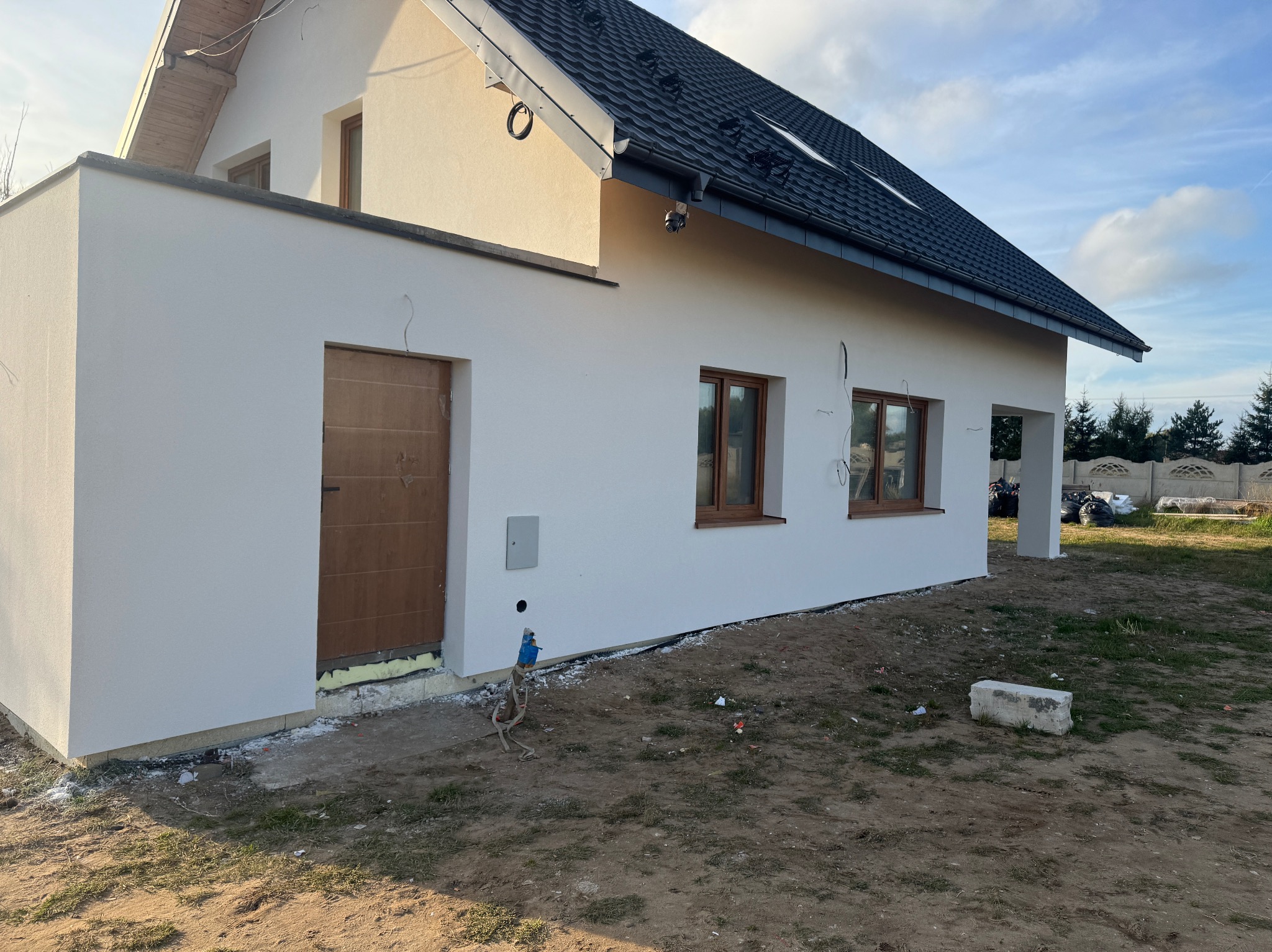 Biała elewacja domu jednorodzinnego z drewnianymi oknami, widok z boku, teren wokół domu jeszcze niezagospodarowany. Nowe orynnowanie.