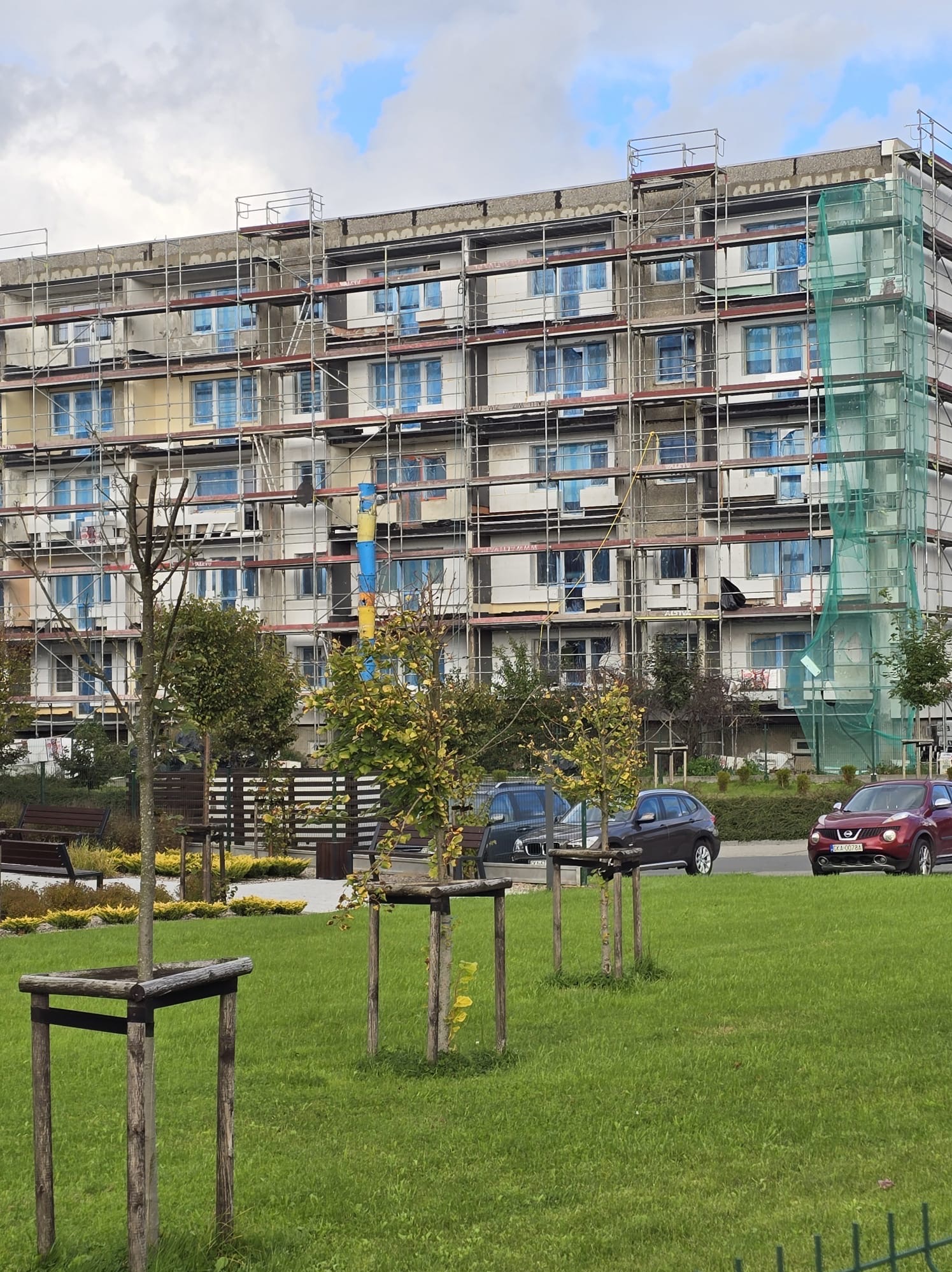 Wielopiętrowy budynek w trakcie ocieplania, osłonięty rusztowaniami i siatką ochronną. Okna zabezpieczone folią. Na pierwszym planie trawnik z młodymi drzewkami.