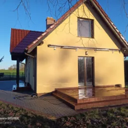 Żółta elewacja domu jednorodzinnego z brązowym dachem, po umyciu, z mokrym tarasem i chodnikiem z kostki brukowej, widoczne kable i osprzęt.