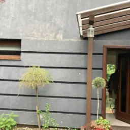 Szara elewacja domu z drewnianą stolarką okienną i drzwiową, ozdobiona roślinnością. Widoczny fragment wejścia z zadaszeniem i lampą.