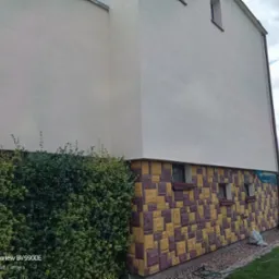 Ściana domu z nową, jasną elewacją i dekoracyjnym wykończeniem imitującym kamień. Widoczny fragment ogrodu z zielonym żywopłotem.