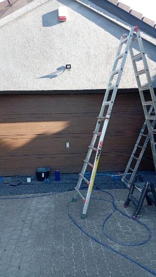 Przygotowania do malowania elewacji: dwie aluminiowe drabiny opierają się o budynek z brązową bramą garażową. Widoczne wiadro i wąż, kostka brukowa.