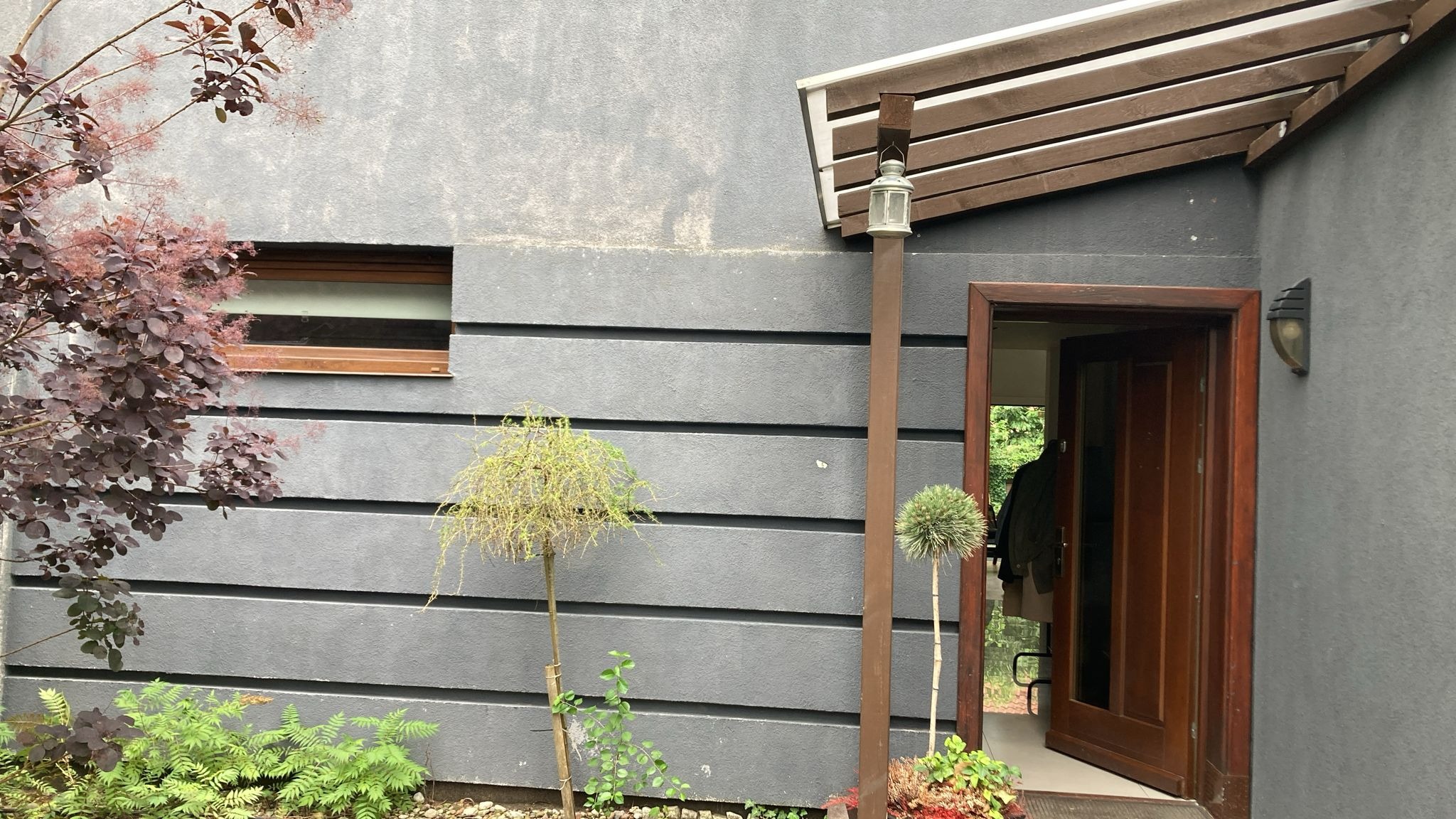 Szara elewacja domu z drewnianą stolarką okienną i drzwiową, ozdobiona roślinnością. Widoczny fragment wejścia z zadaszeniem i lampą.