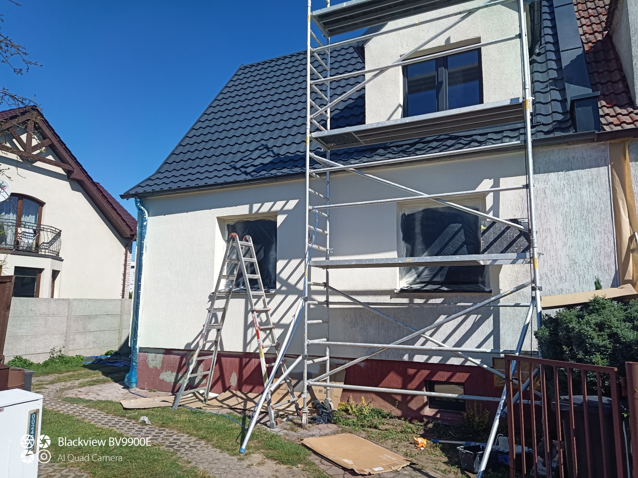 Biała elewacja domu w trakcie prac, z rusztowaniem i drabiną. Okna zabezpieczone folią. Widoczne przygotowania do malowania, w słoneczny dzień.