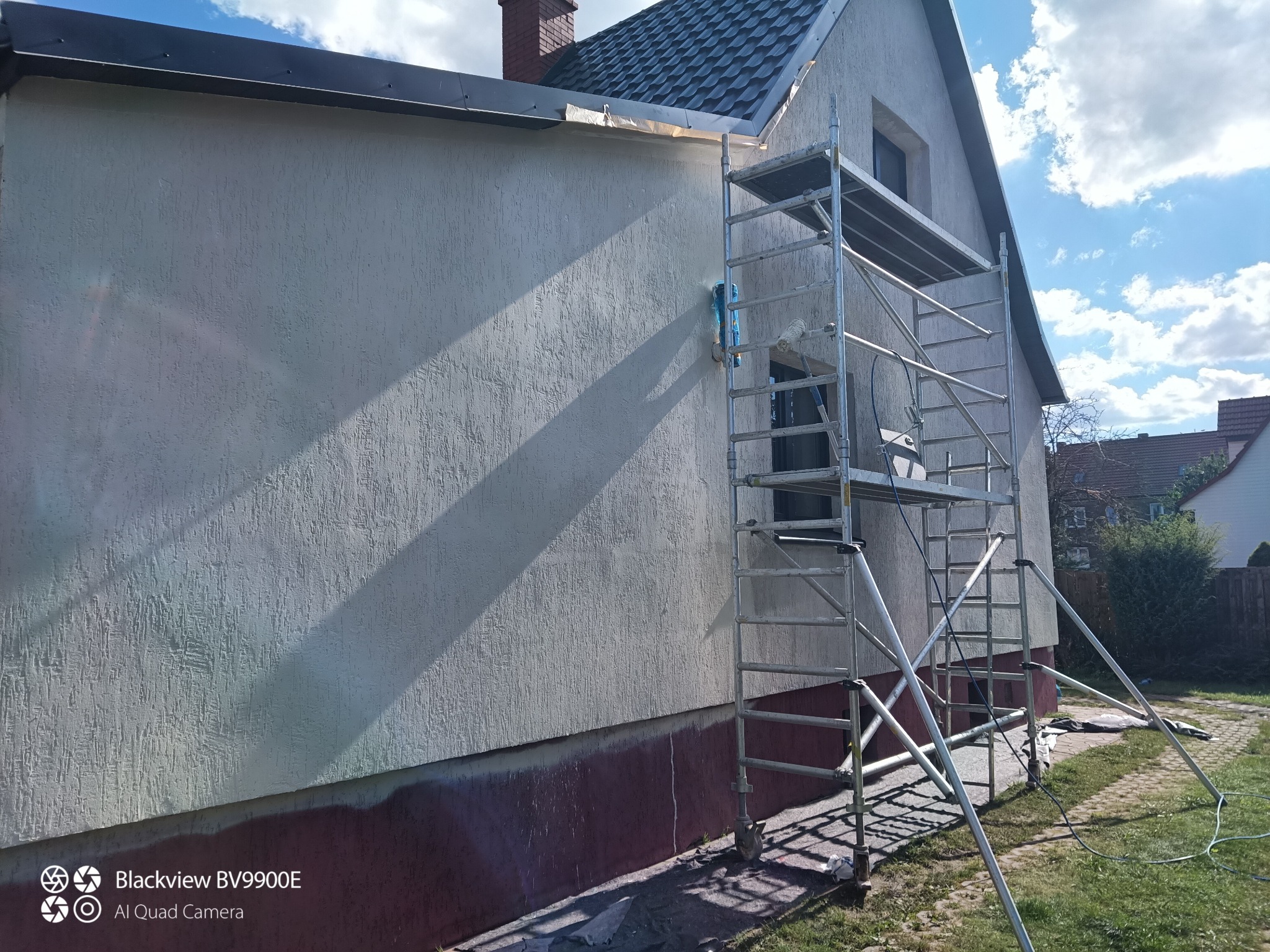 Elewacja domu w trakcie prac: pomalowana górna część, rusztowanie z rolką i pędzlem. Widoczne niedomalowane fragmenty przy cokole. Słoneczny dzień.