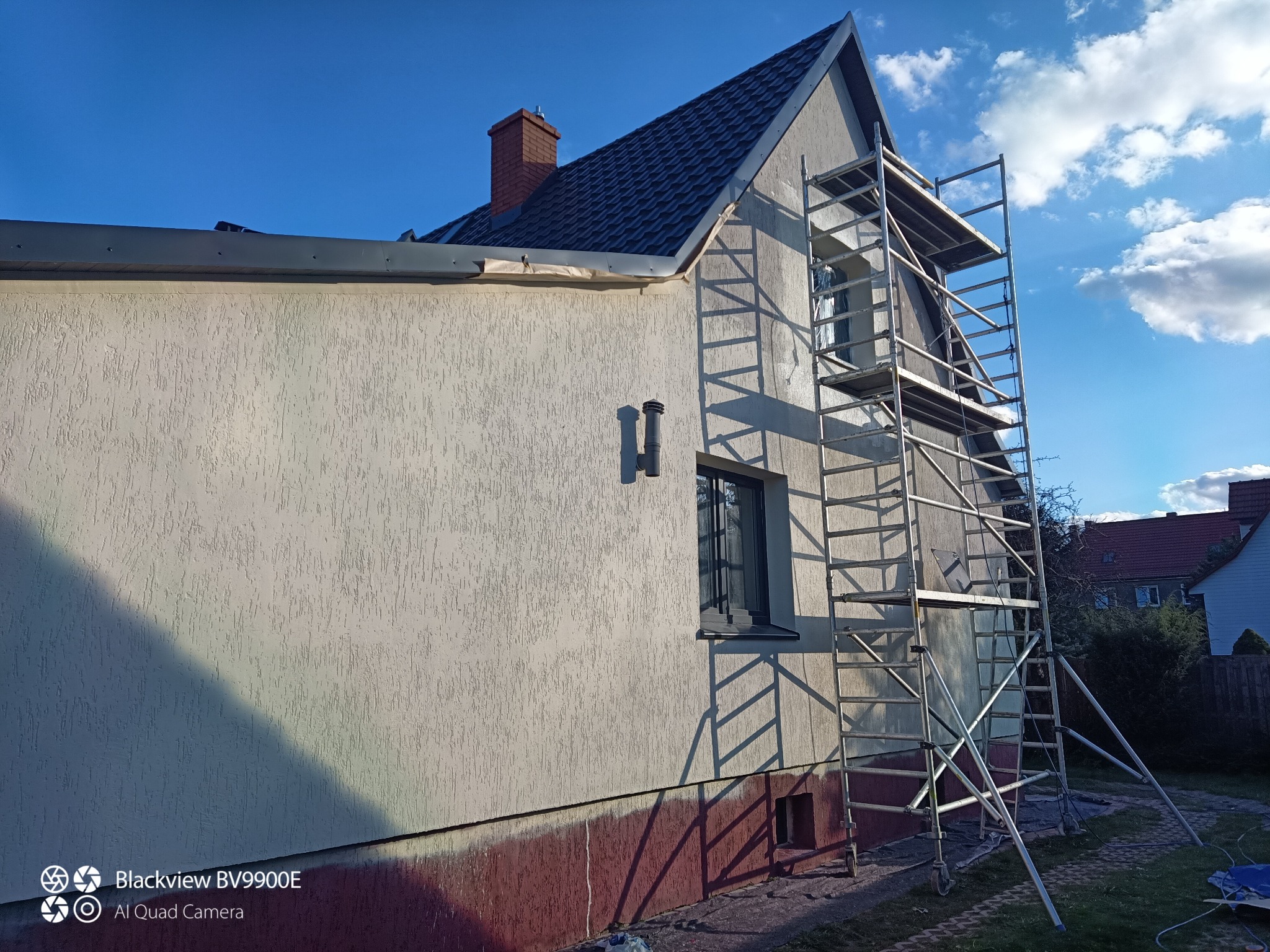 Elewacja domu w trakcie malowania, z widocznym rusztowaniem i częściowo pomalowaną dolną partią. Słoneczny dzień, błękitne niebo.