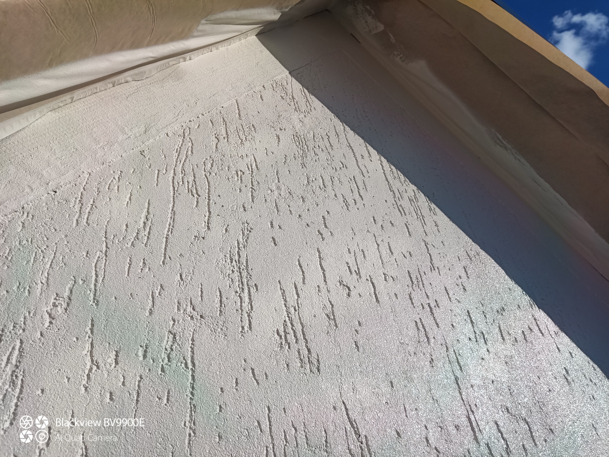 Biała elewacja budynku z fakturą 'baranka', zabezpieczona folią malarską u góry, widok z dołu pod kątem. Fragment nieba w prawym górnym rogu.