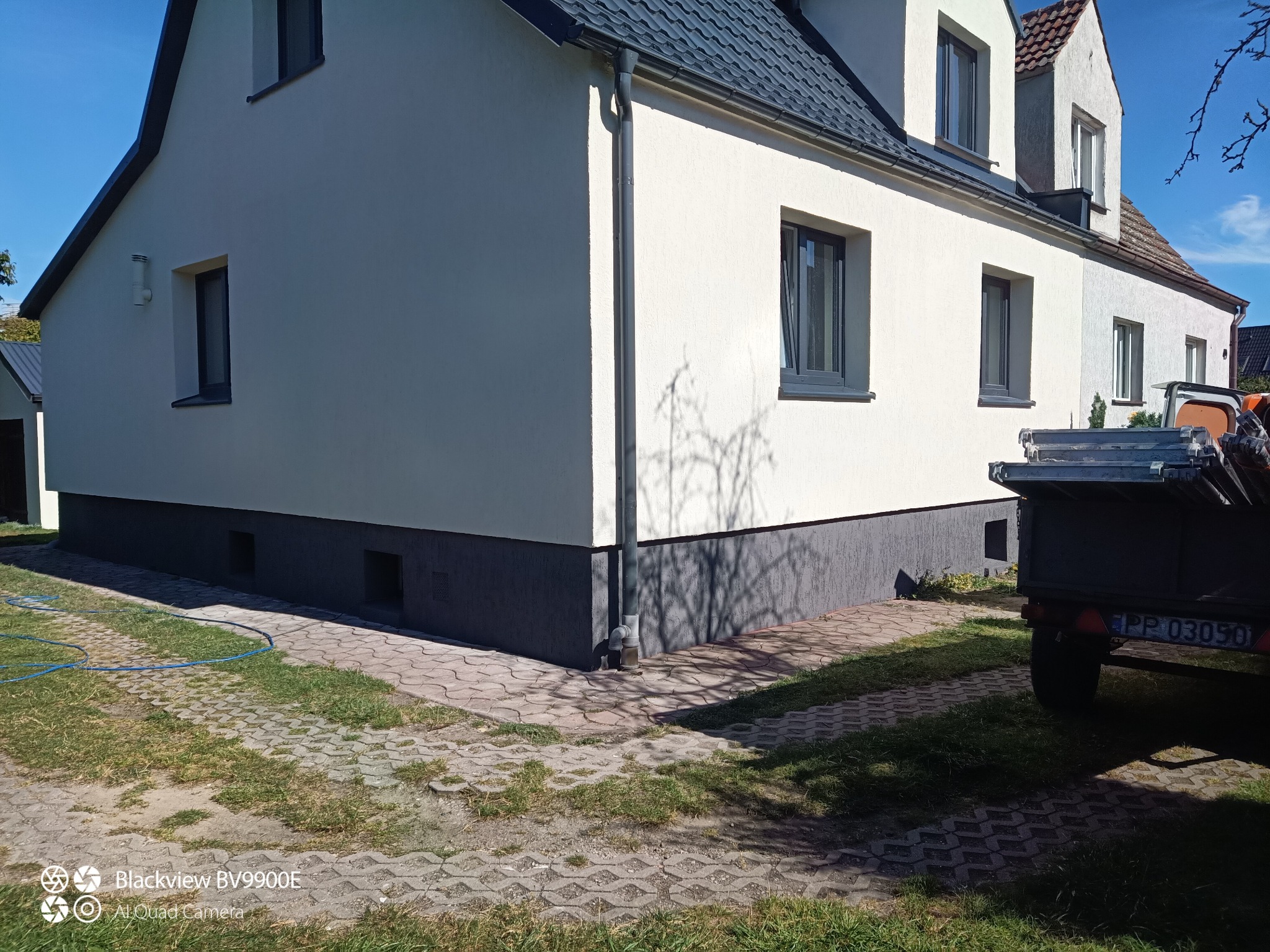 Odnowiona elewacja domu w Trzciance z szarą podmurówką i białymi ścianami. Widoczne okna z szarymi ramami i fragment przyczepki.