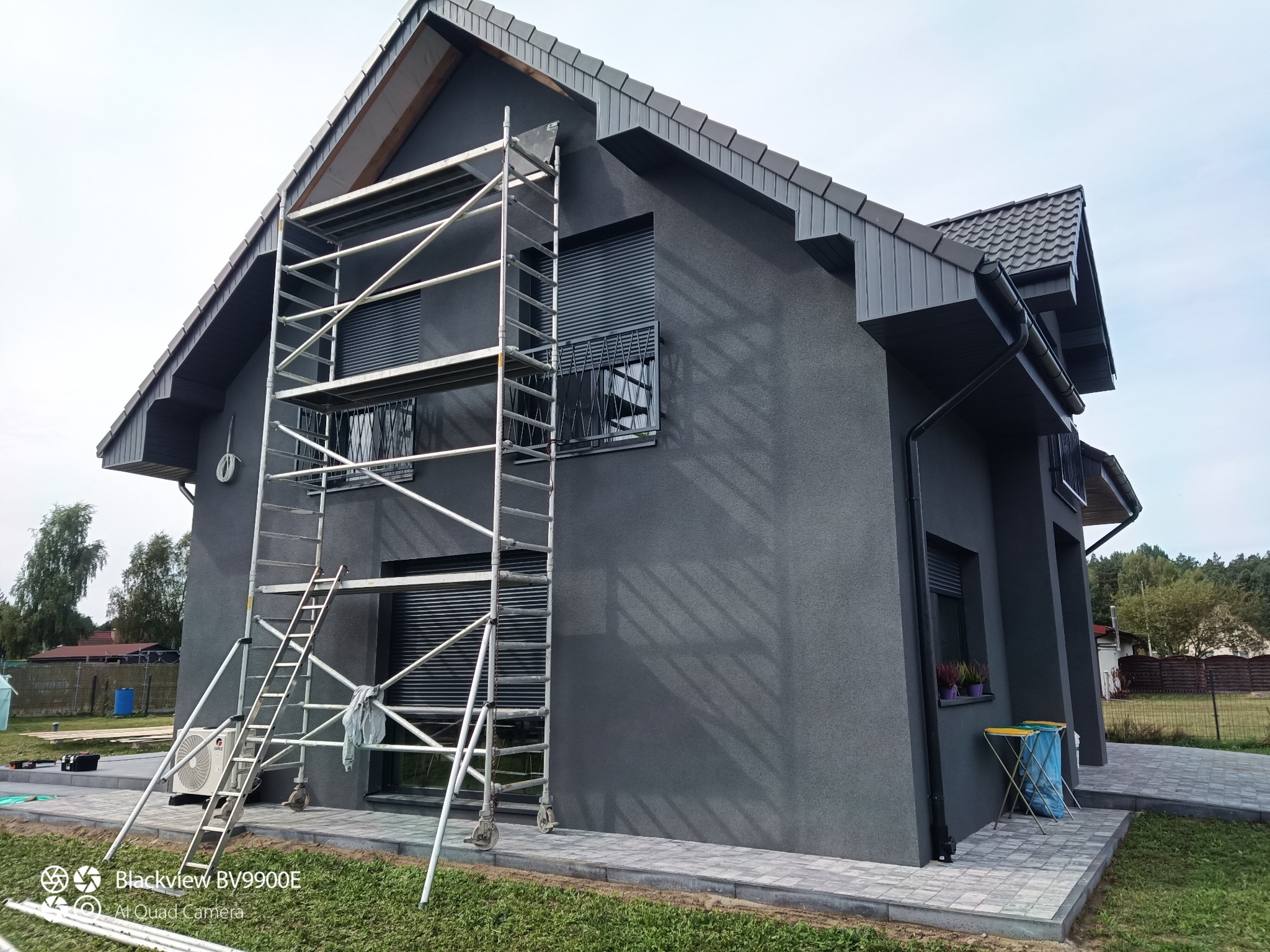 Elewacja domu pomalowana na grafitowo, widoczne rusztowanie przy ścianie, okna z żaluzjami zewnętrznymi, kostka brukowa wokół domu.