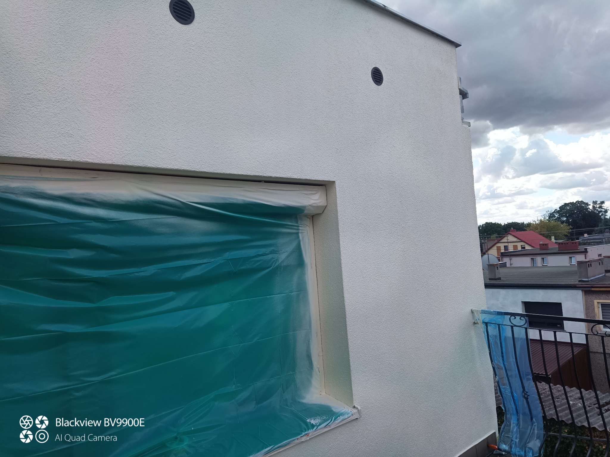 Biała elewacja domu w trakcie prac, okno zabezpieczone folią malarską. Widoczne detale faktury tynku i elementy wentylacyjne. Częściowy widok balkonu z metalową barierką.