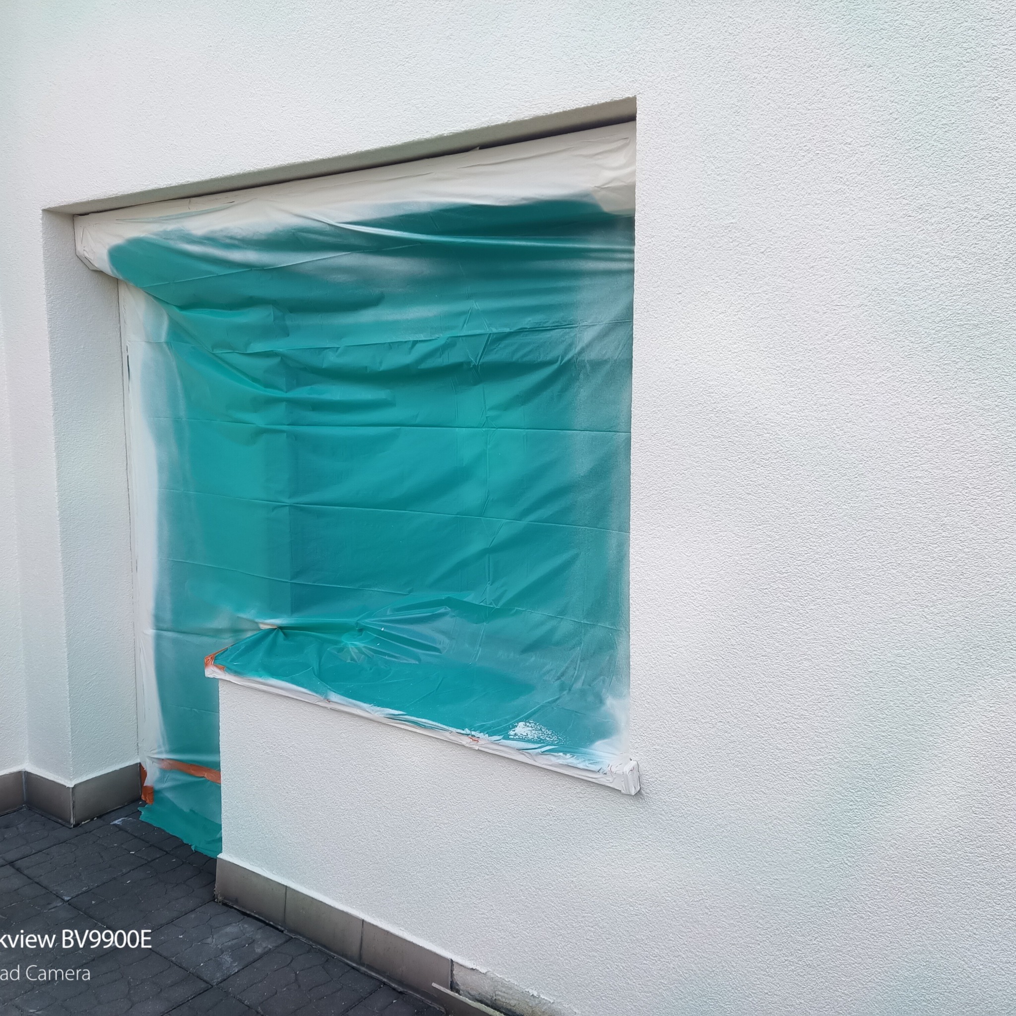 Biała elewacja budynku z oknem zabezpieczonym folią malarską. Przygotowanie do prac malarskich na zewnątrz. Widoczna faktura tynku.