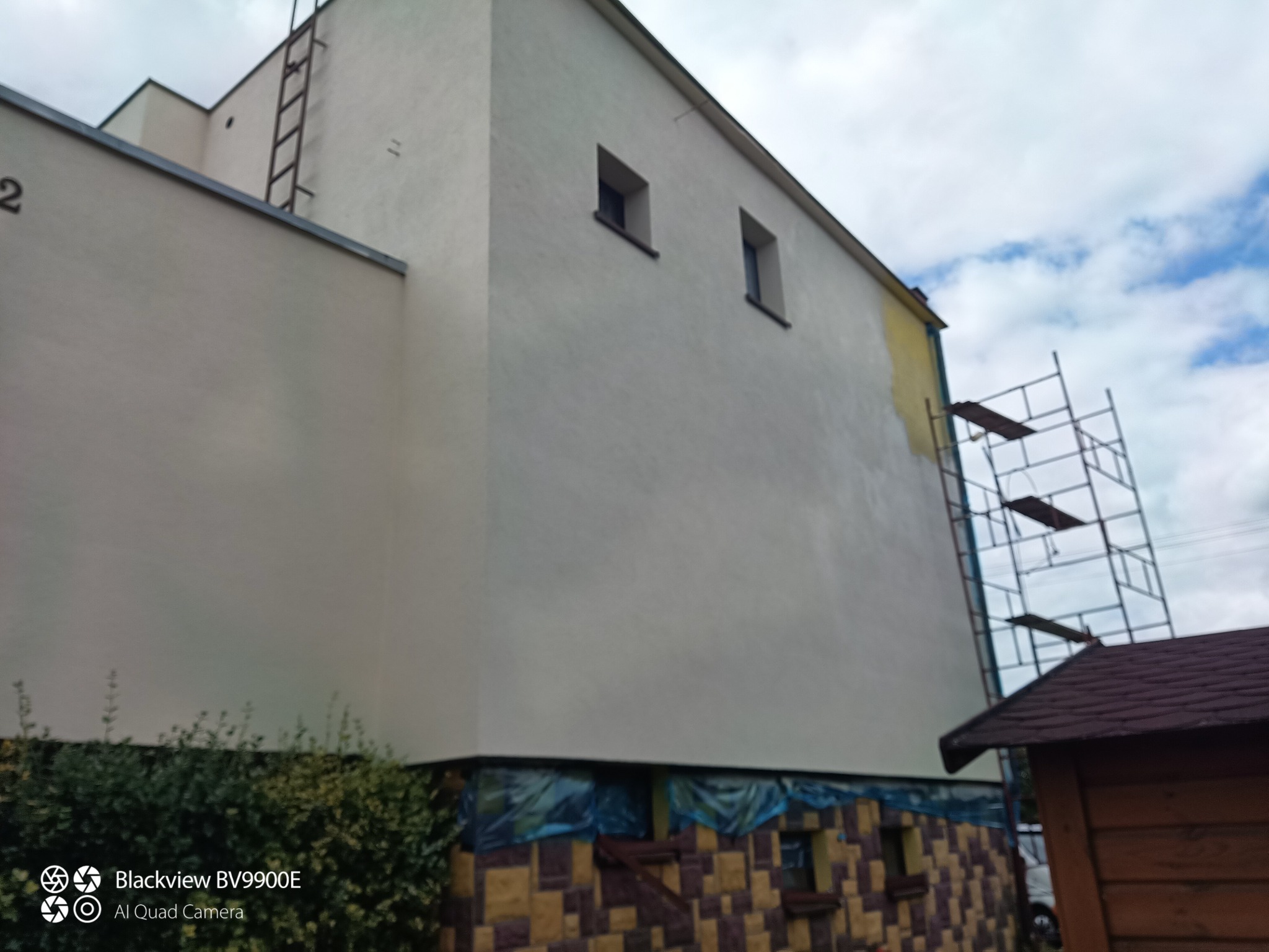 Ściana budynku w trakcie malowania, widoczne rusztowanie i częściowo pomalowana elewacja na żółto, zabezpieczone okna folią.