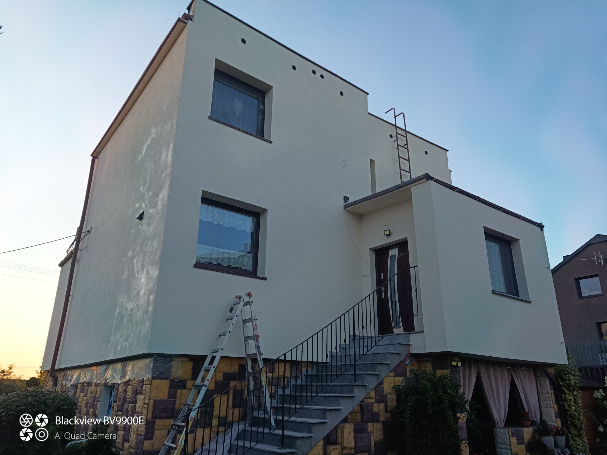 Pomalowana na kremowo elewacja domu z drabinką na ścianie i schodami wejściowymi. Widoczne okna z firankami i kamienna podmurówka.