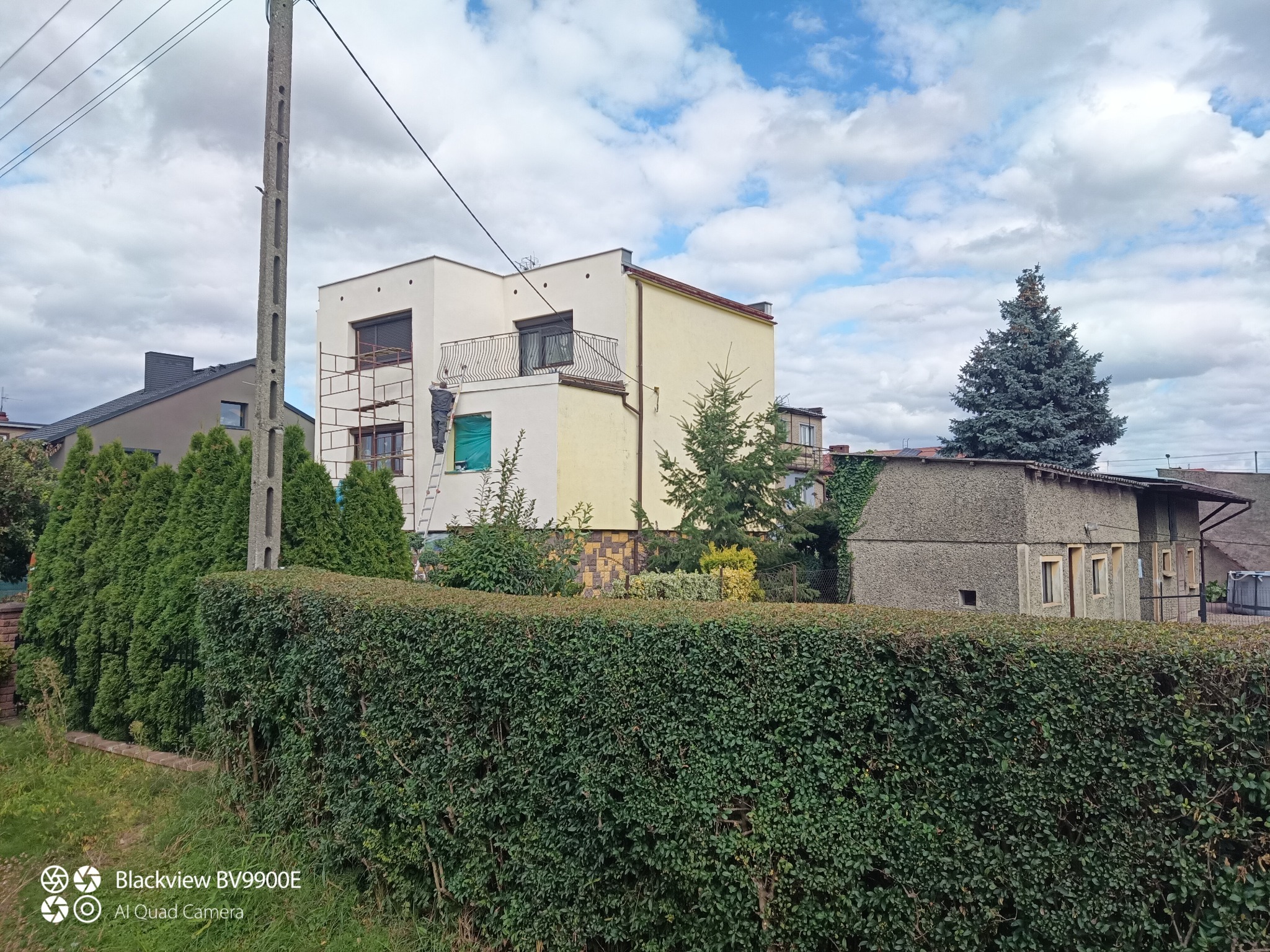 Pracownik na drabinie maluje fragment elewacji dwukondygnacyjnego domu. Widoczne rusztowanie, zieleń i błękitne niebo z chmurami w tle. Prace na zewnątrz budynku.