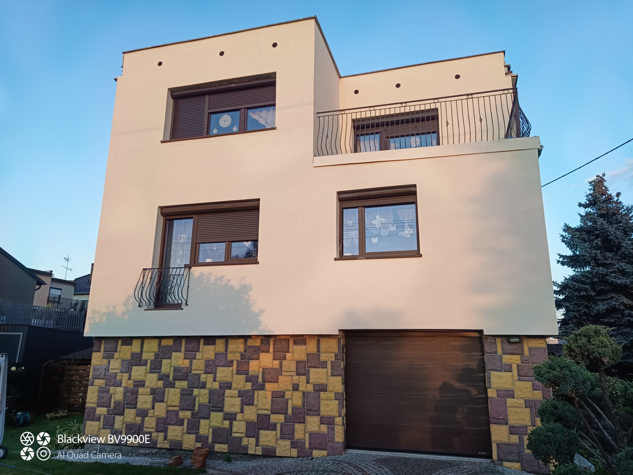 Dwupiętrowy dom z jasną elewacją i balkonem, z garażem. Dolna część budynku wykończona dekoracyjną okładziną w odcieniach żółci i brązu. Widok z poziomu gruntu.