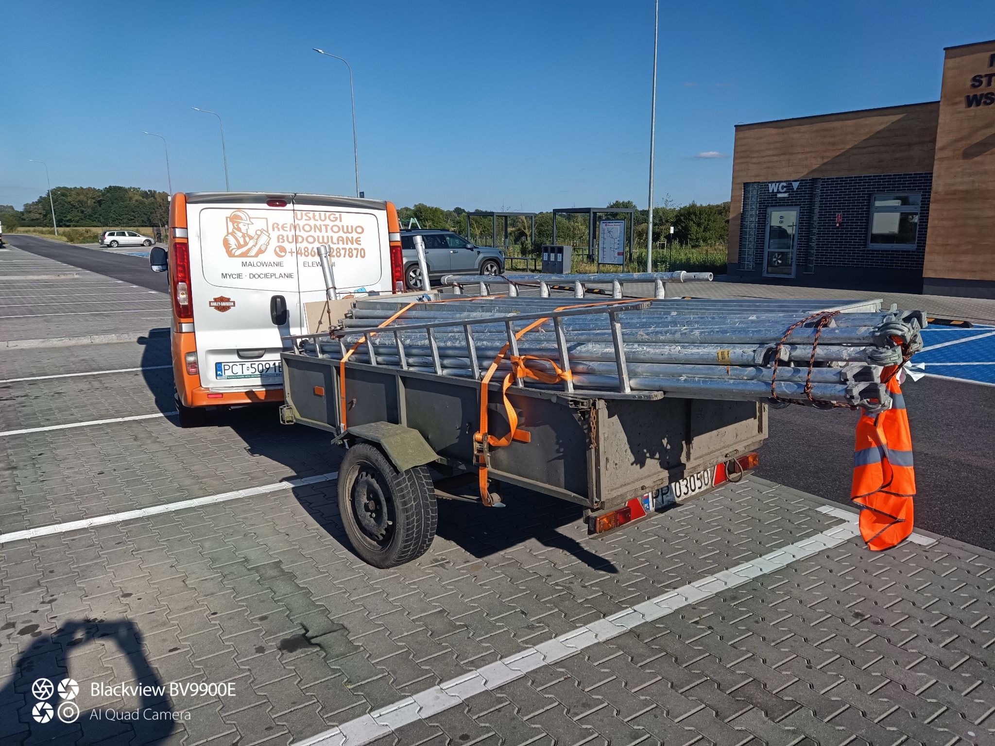 Przyczepka z rusztowaniem, zaparkowana obok samochodu firmy remontowej. Z tyłu budynek z toaletami. Słoneczny dzień, błękitne niebo.