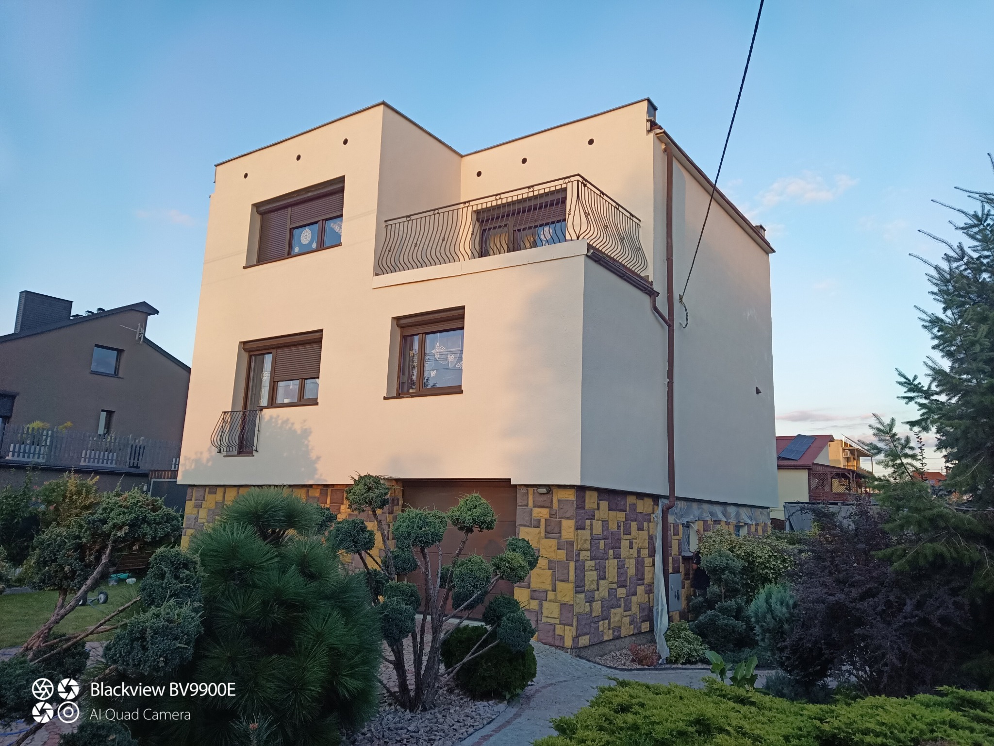 Nowoczesny dom z beżową elewacją, balkonem z ozdobną balustradą i kamienną podmurówką w odcieniach żółci i brązu. Uwagę zwraca starannie wypielęgnowany ogród.