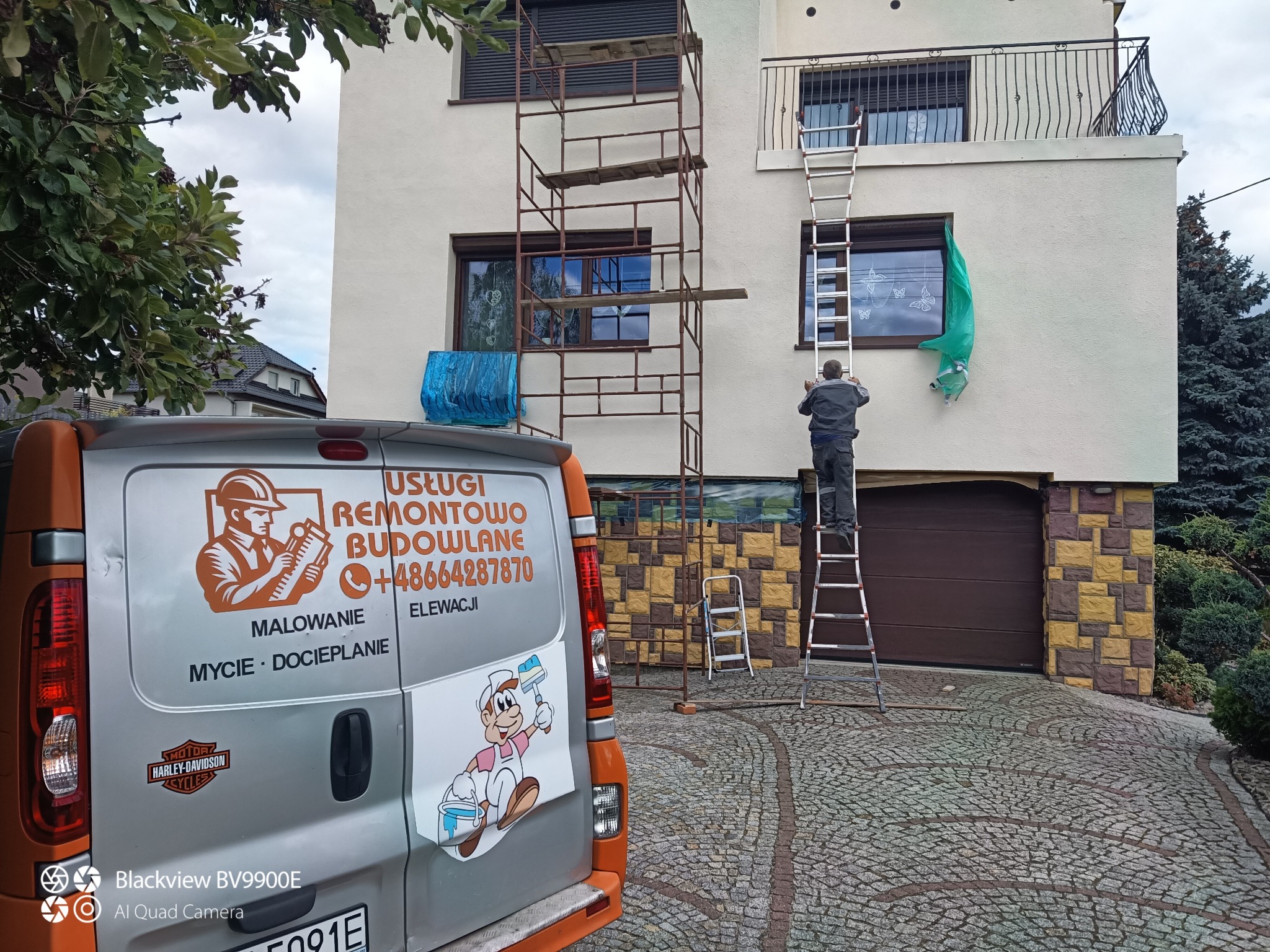 Pracownik na drabinie, maluje elewację domu. Widoczne rusztowanie, okna zabezpieczone folią. Obok zaparkowany bus z reklamą usług remontowo-budowlanych.
