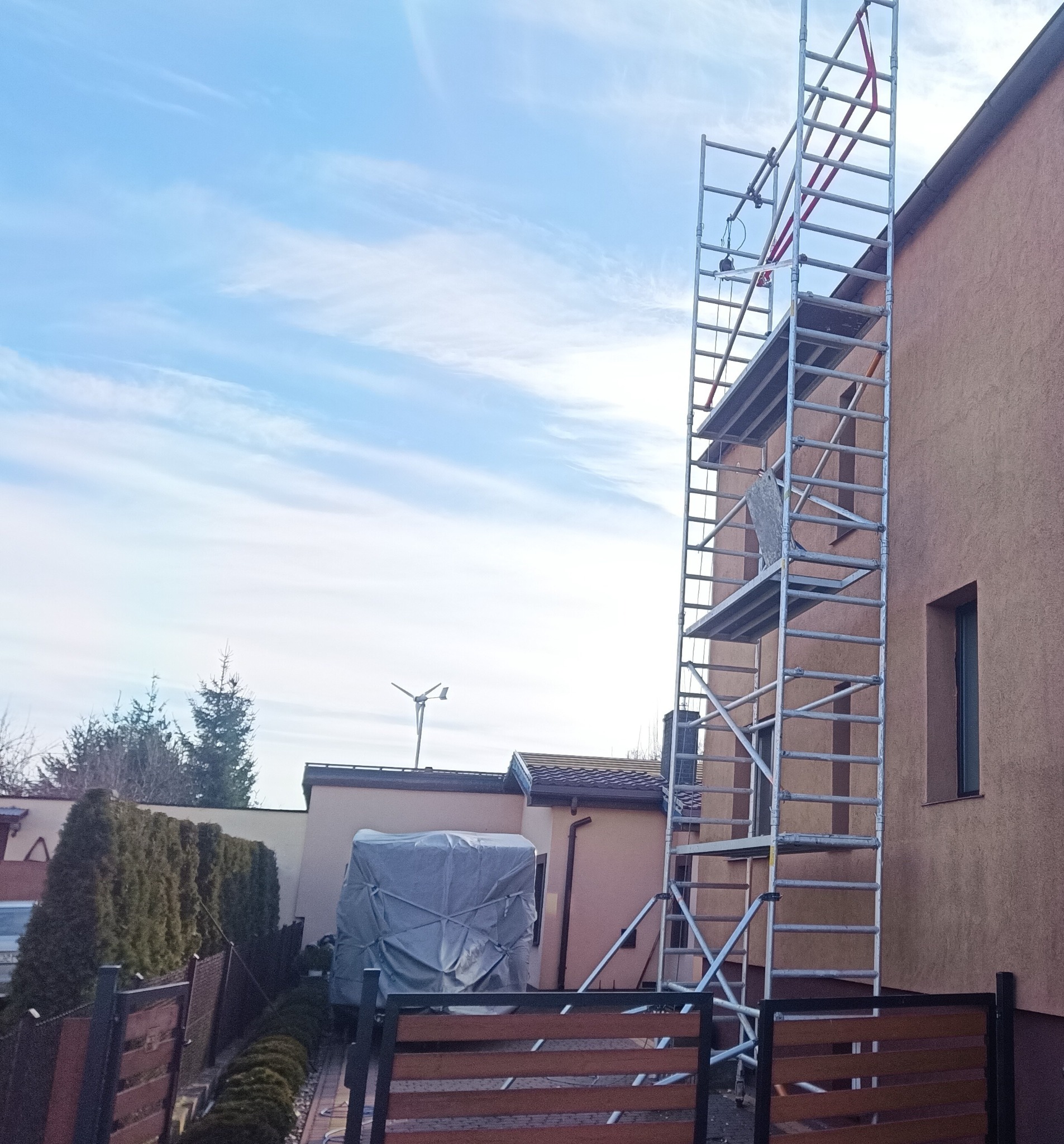 Elewacja domu w trakcie prac. Widoczne rusztowanie aluminiowe, fragment pomalowanej ściany w odcieniach brązu, błękitne niebo. Prace na wysokości.