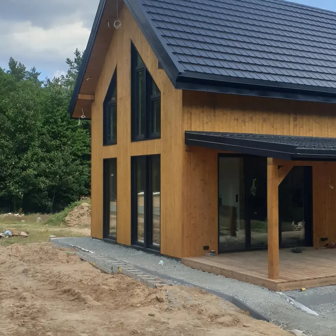 Surowy, nowoczesny dom z drewna z czarną dachówką i dużymi oknami, na etapie wykończenia terenu wokół budynku.