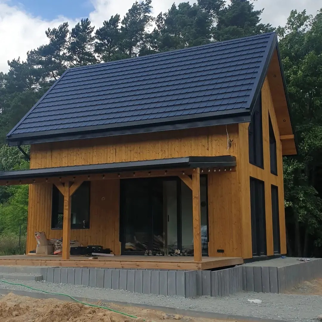 Nowoczesny, parterowy dom z drewna z ciemną, metalową dachówką, widoczne elementy konstrukcyjne i drewniana podłoga tarasu w trakcie budowy, zarysowane elementy podmurówki.