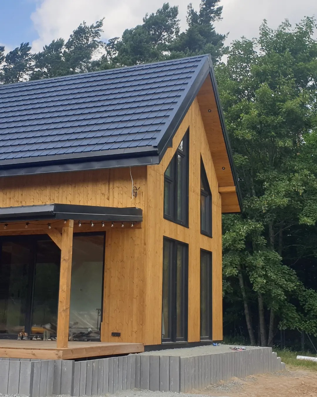 Nowoczesny dom szkieletowy z ciemną dachówką i dużymi oknami, widok z zewnątrz, częściowo wykończony taras z szarych bloczków.