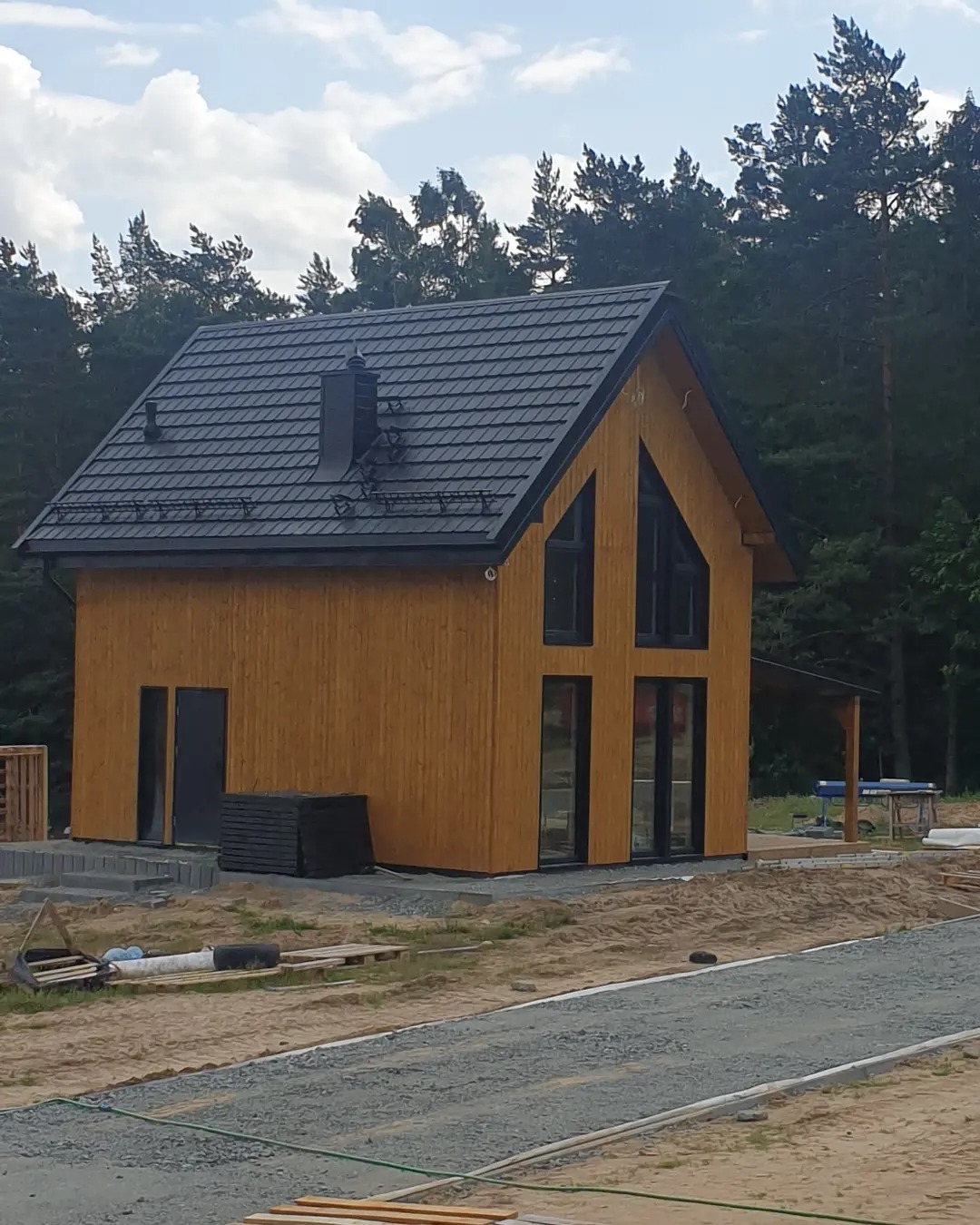 Nowoczesny dom z drewnianą elewacją i czarną dachówką w trakcie budowy, widoczne materiały budowlane i teren przygotowany pod zagospodarowanie.