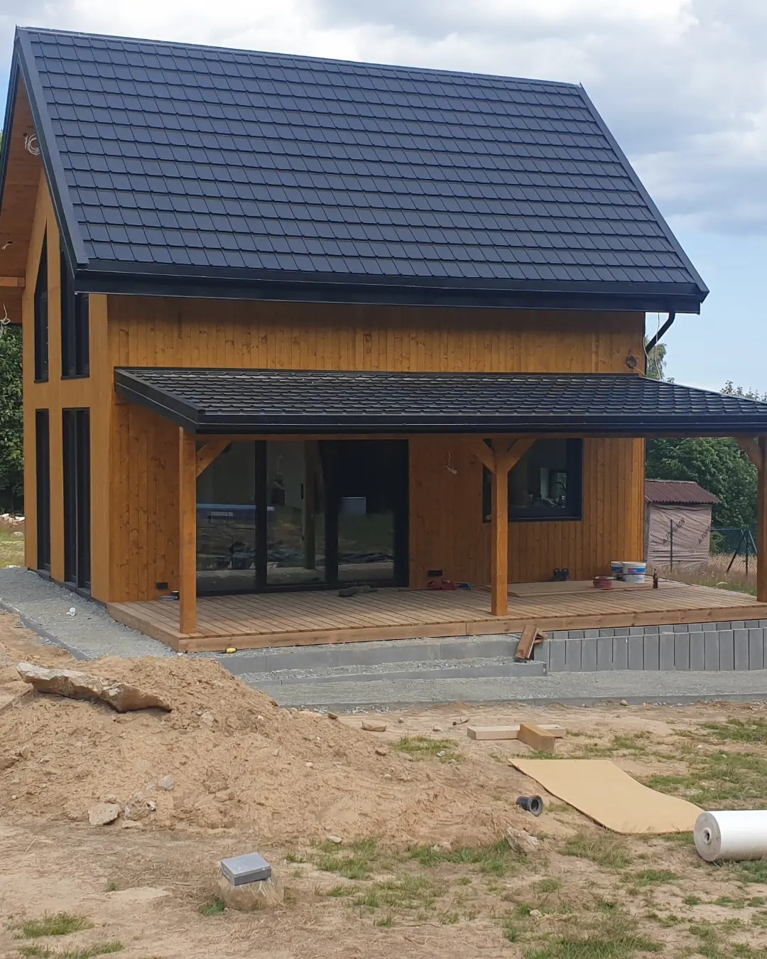 Nowoczesny, dwukondygnacyjny dom z drewnianą elewacją i ciemnym dachem, z zadaszonym tarasem i widocznymi elementami wykończeniowymi na zewnątrz.