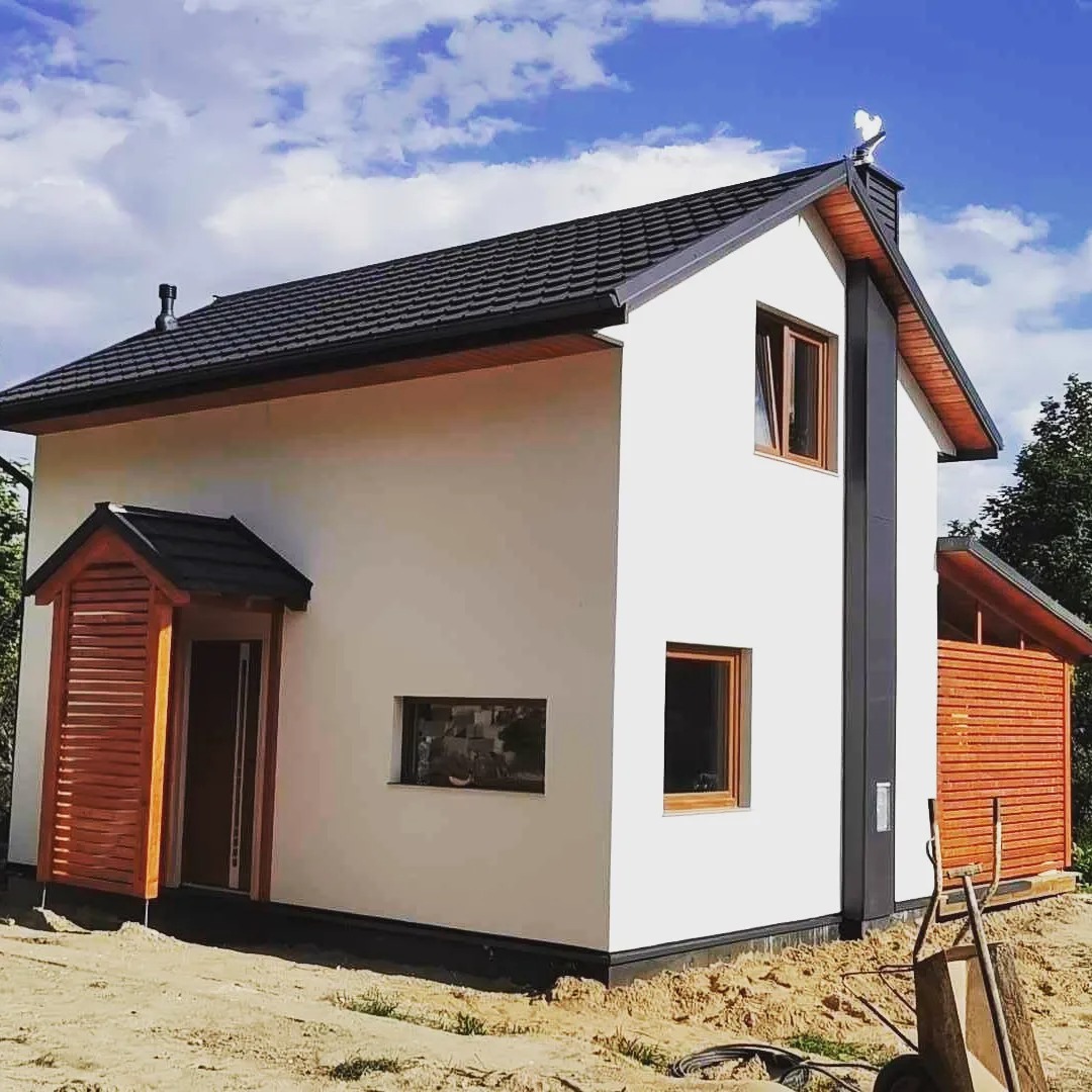 Nowoczesny, dwukondygnacyjny dom z białymi ścianami, grafitowym dachem i drewnianymi elementami wykończeniowymi, widoczny wóz budowlany na niezagospodarowanym terenie.