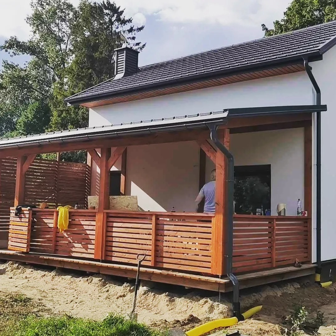 Nowoczesny dom z drewnianą werandą w trakcie budowy, widoczne narzędzia i materiały budowlane, częściowo wykończona elewacja i teren wokół.