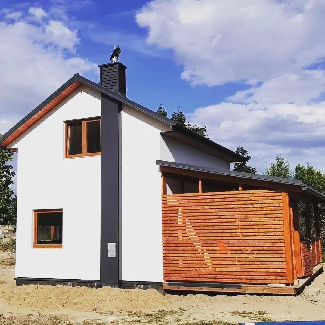 Nowoczesny dom szkieletowy z białymi ścianami, drewnianą elewacją i ozdobną figurką ptaka na kominie, na tle błękitnego nieba.