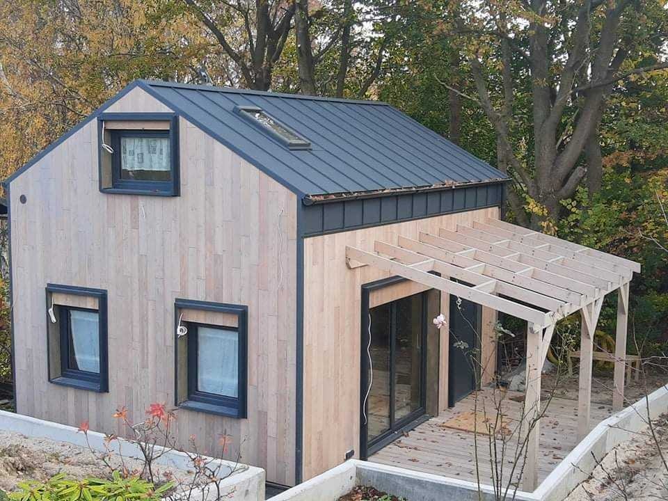 Nowoczesny dom z drewnianą elewacją i metalowym dachem w trakcie budowy z widoczną konstrukcją drewnianej pergoli, okna z roletami zewnętrznymi.