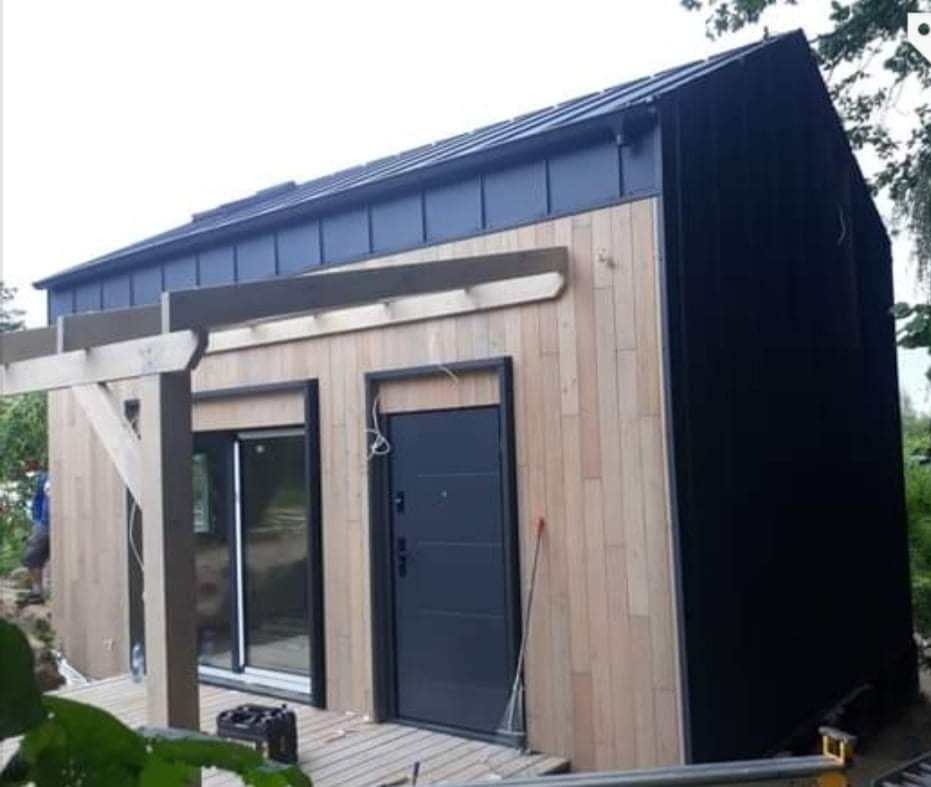 Nowoczesny, mały budynek z drewnianą elewacją i ciemnym dachem, widoczna drewniana pergola w trakcie budowy, z czarnymi elementami wykończeniowymi.