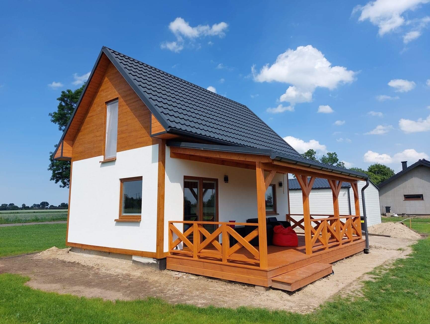 Nowoczesny, parterowy dom z drewnianą elewacją na poddaszu i przestronnym, zadaszonym tarasem z drewnianą balustradą w kształcie litery X, na tle błękitnego nieba z obłokami.