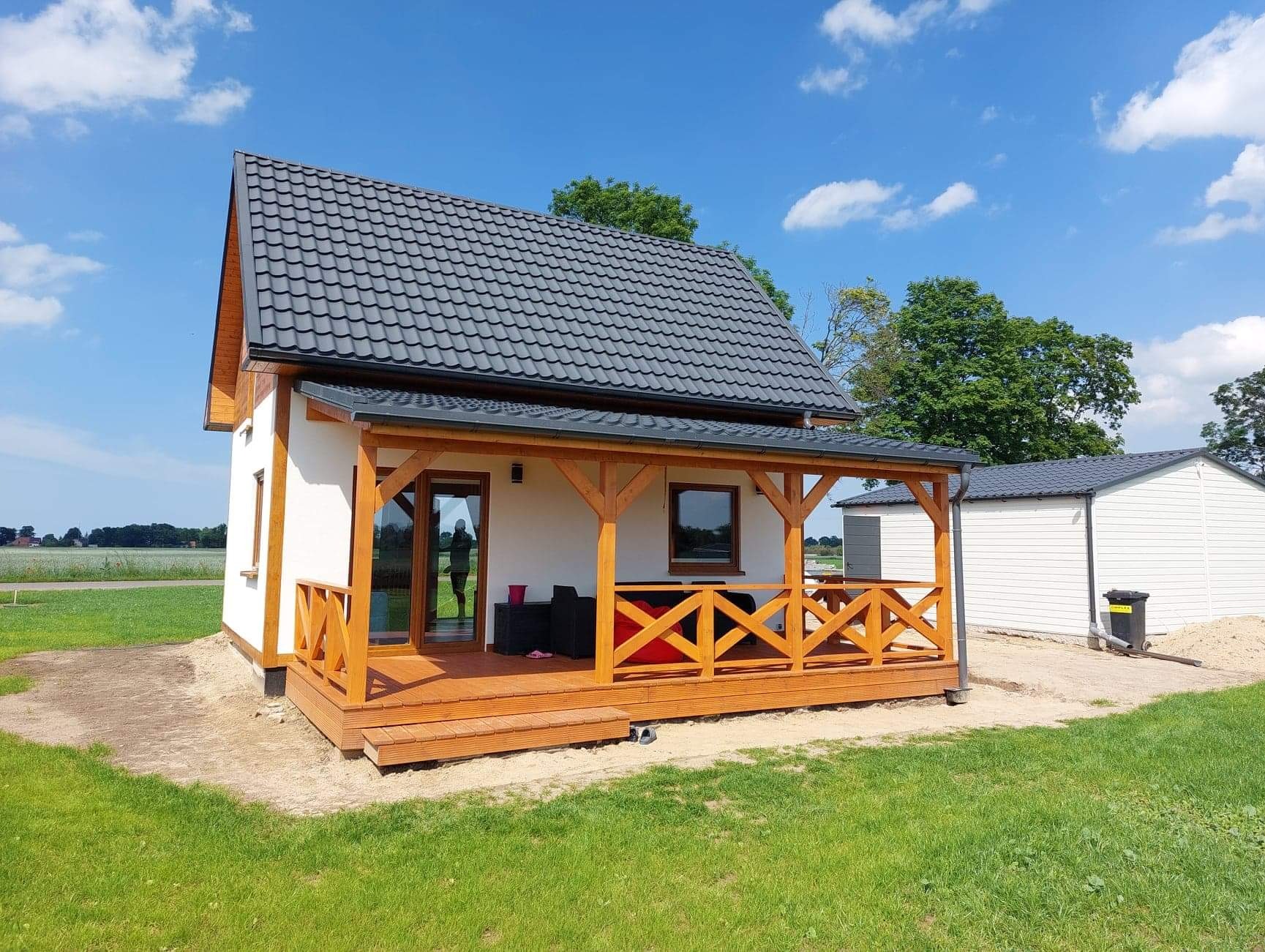 Niewielki, parterowy dom z drewnianą werandą i grafitową dachówką, otoczony zielonym trawnikiem i błękitnym niebem z delikatnymi chmurami. W oknie widoczna odbijająca się sylwetka osoby.